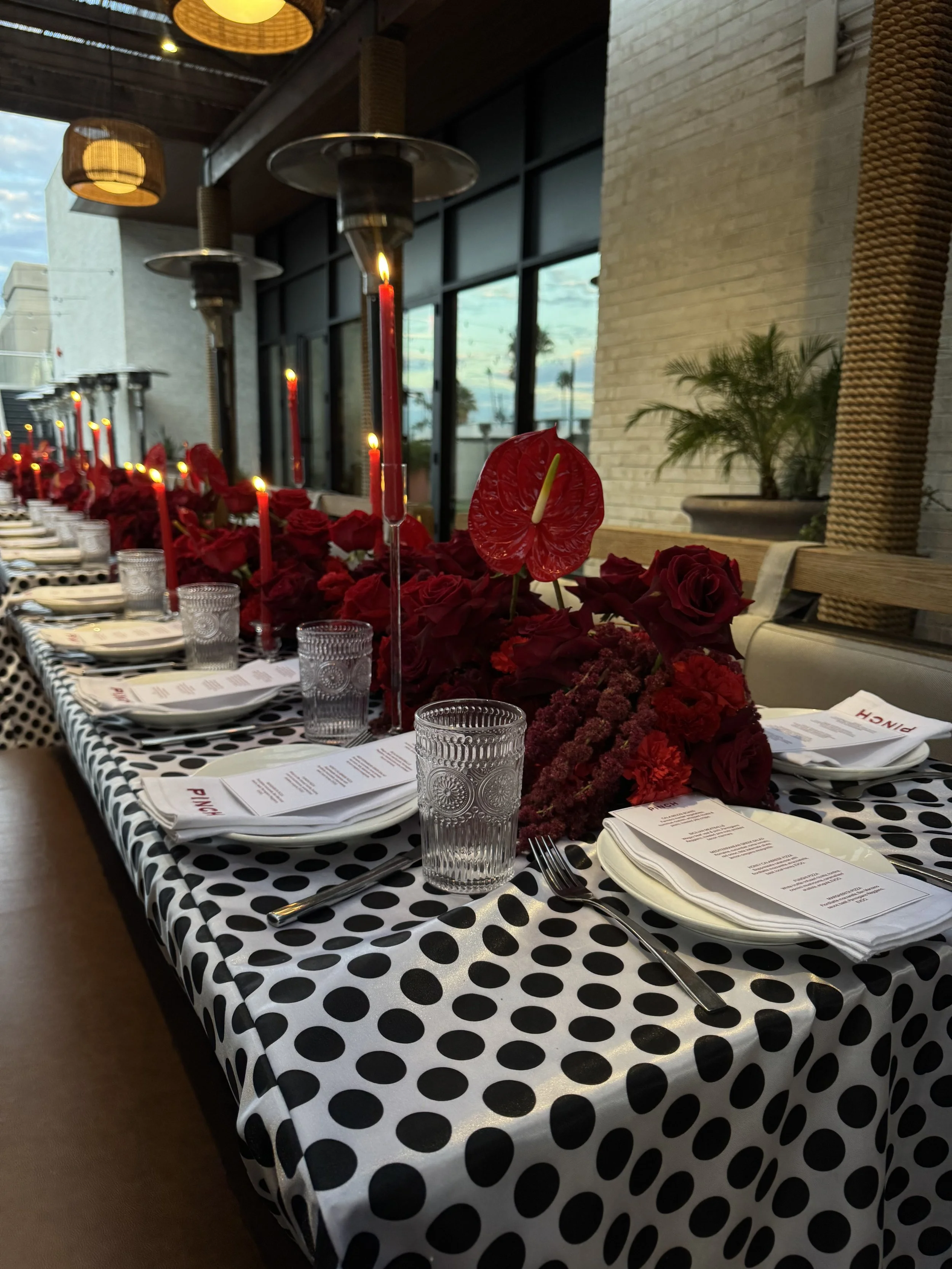 A dinner table set with black and white polka dot tablecloth, red flowers, red candles, drinking glasses, and menus, in a modern restaurant with large windows and hanging lamps.