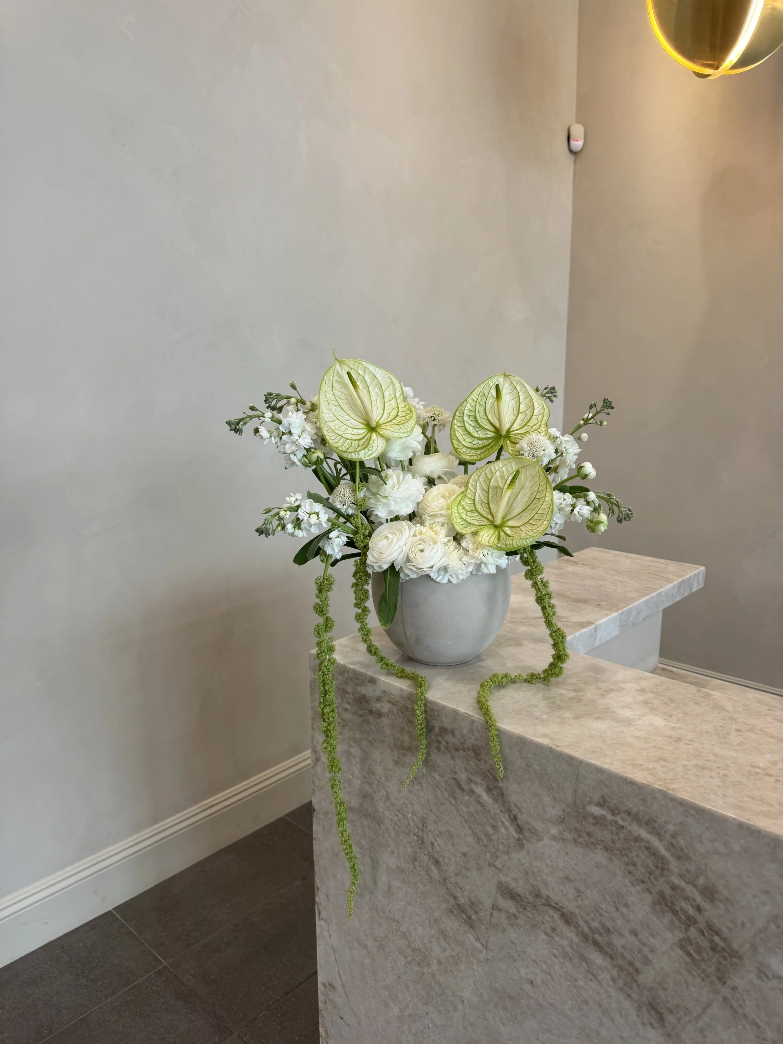 A bouquet of white flowers with green anthuriums in a white vase on a beige marble table against a plain light-colored wall.