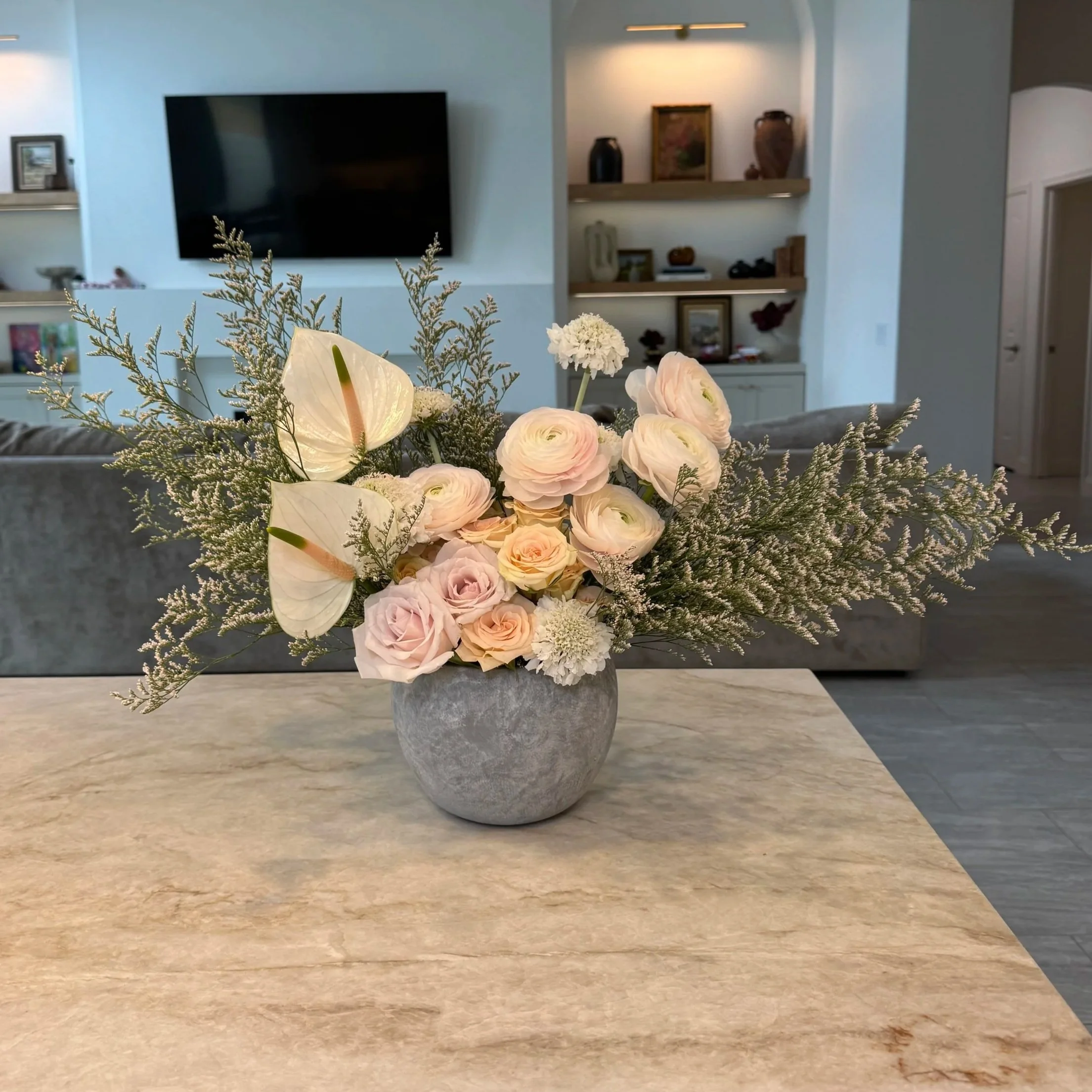 Close-up of a bouquet of light pink roses, white anthuriums, and assorted greenery in a gray stone vase on a beige countertop.