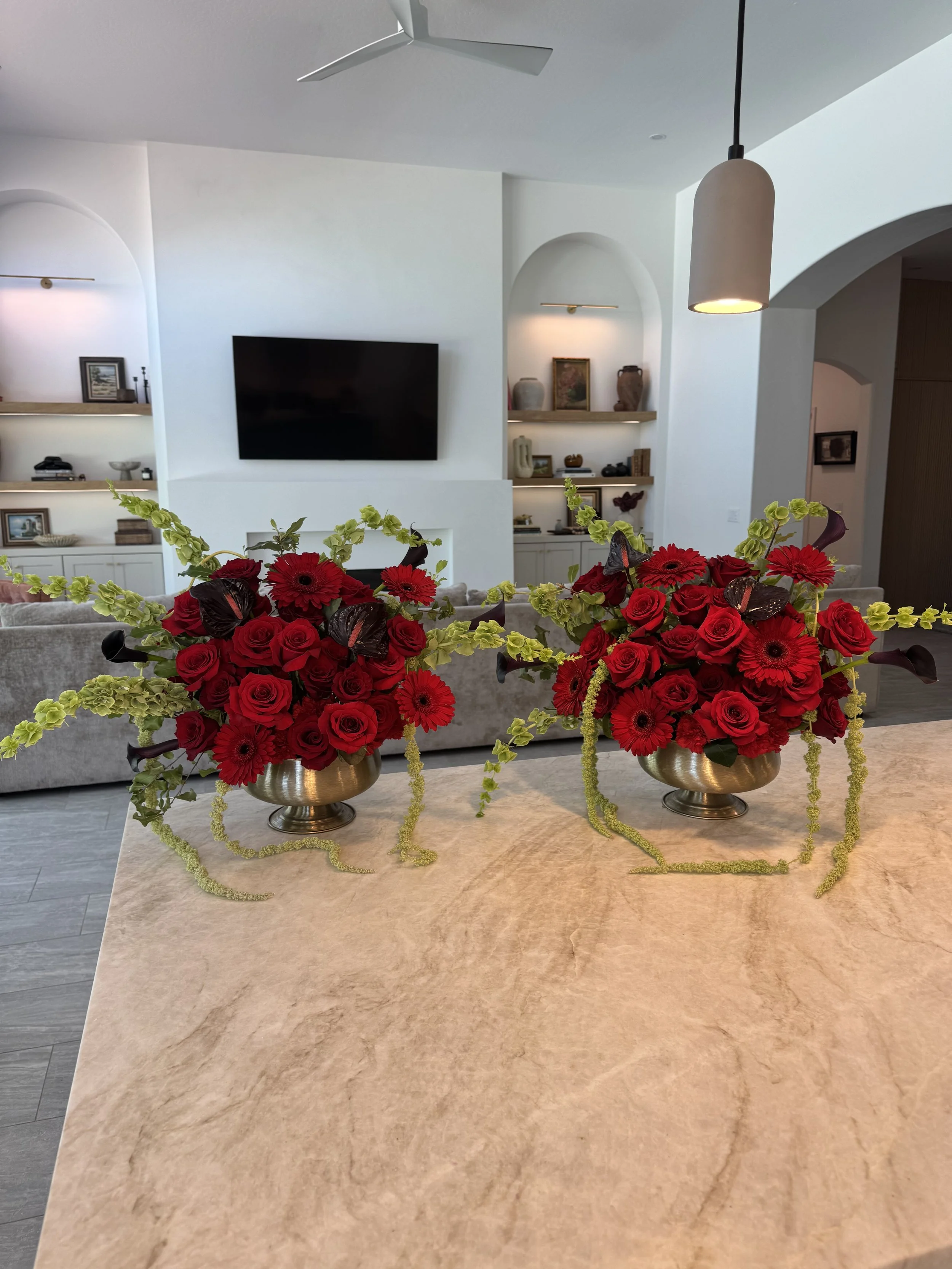 A beige marble kitchen island with three silver vases filled with red roses, green hydrangeas, and dark calla lilies, and gold beaded accents.