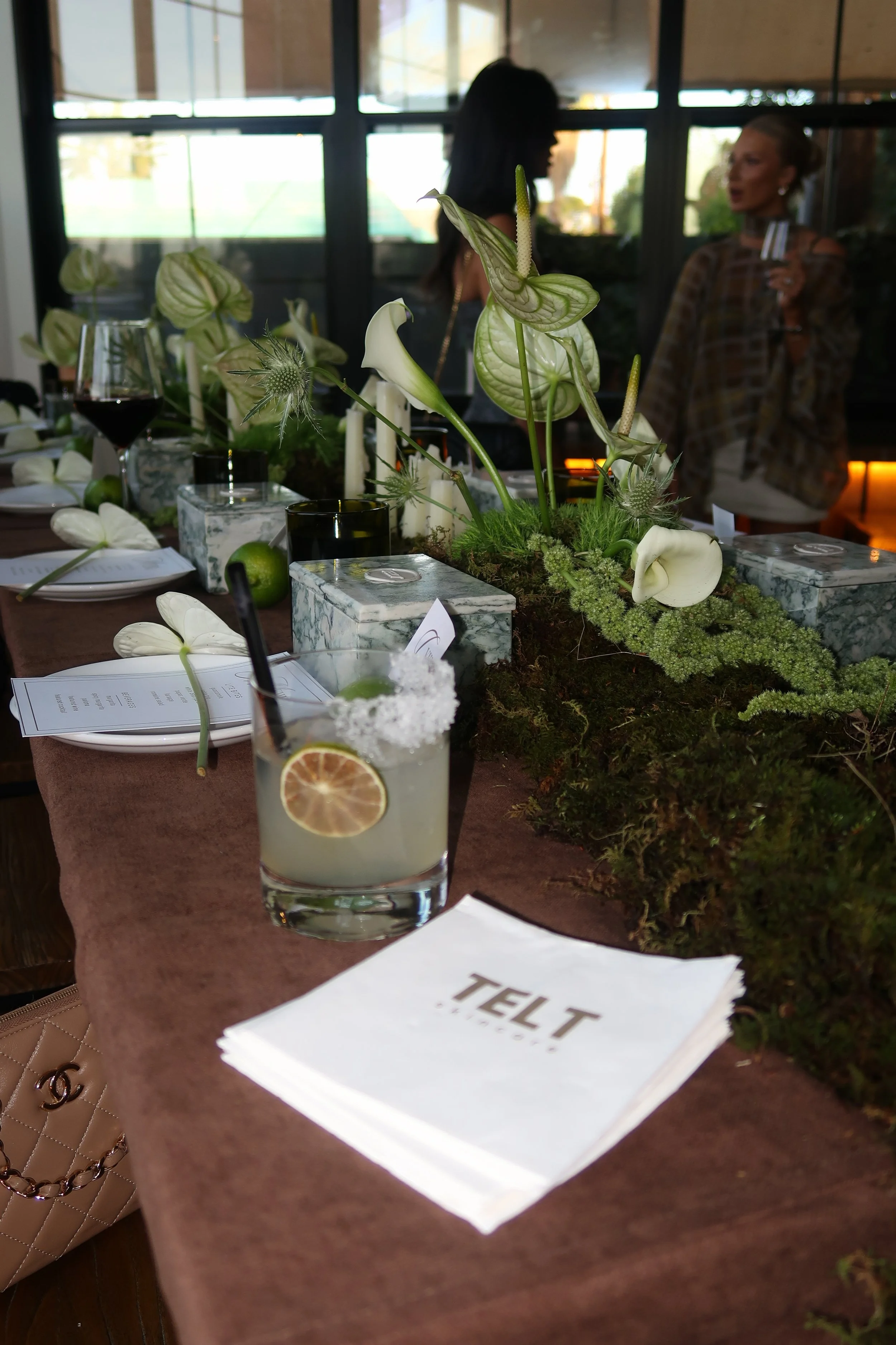 A dining table with floral decorations, candles, wine glasses, and a cocktail with a lemon slice. There are people in the background behind a glass window, one woman holding a drink.