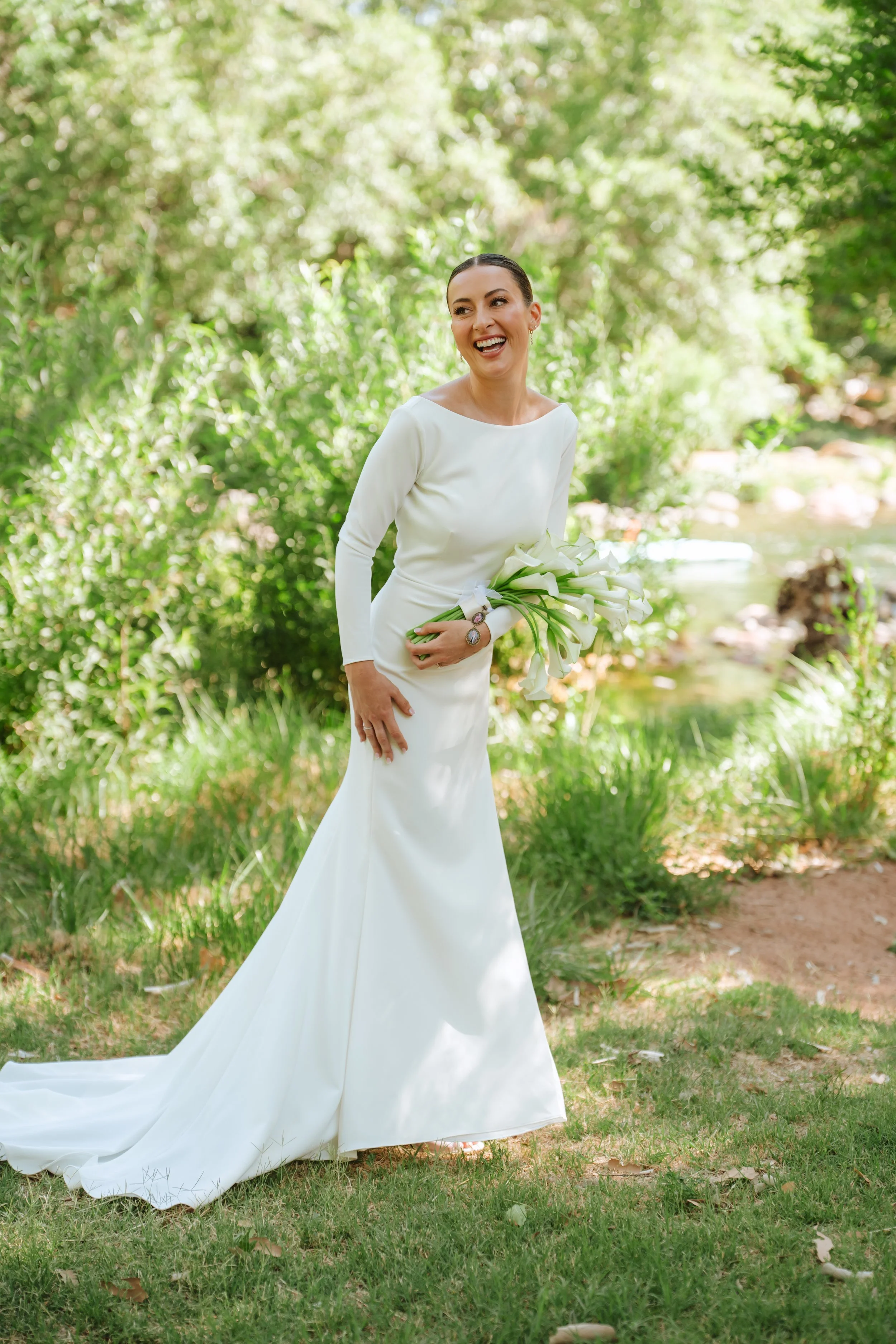 A woman in a white gown holding a bouquet of white flowers standing outdoors in a lush green setting with trees and grass.