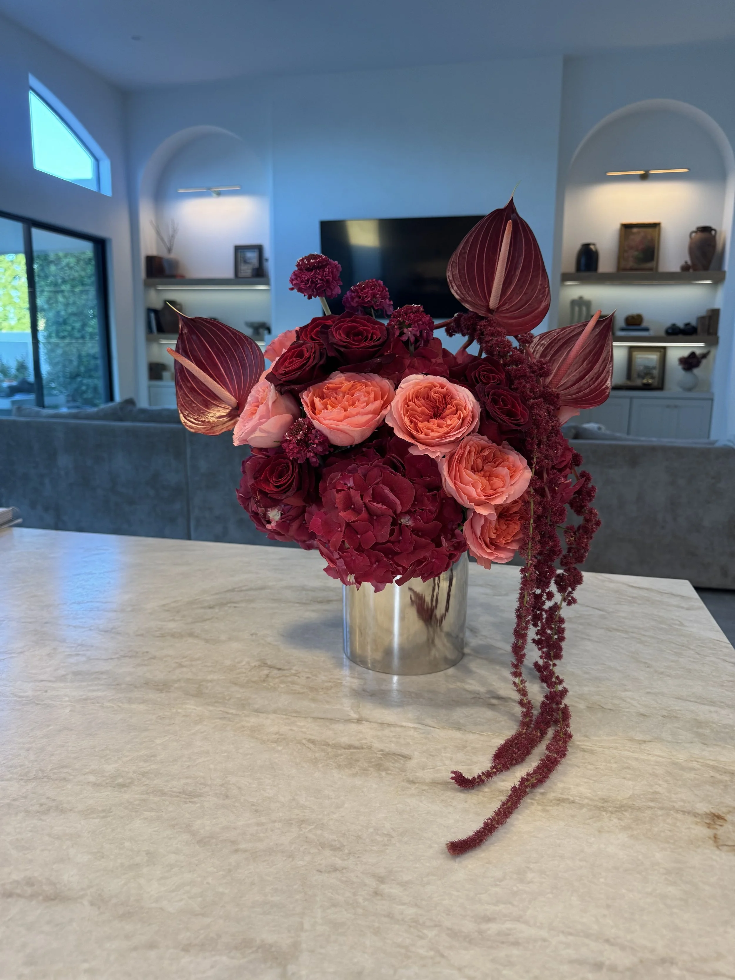 A bouquet of pink, red, and purple flowers in a metallic vase on a light-colored table in a living room with shelves, artwork, and a large television in the background.