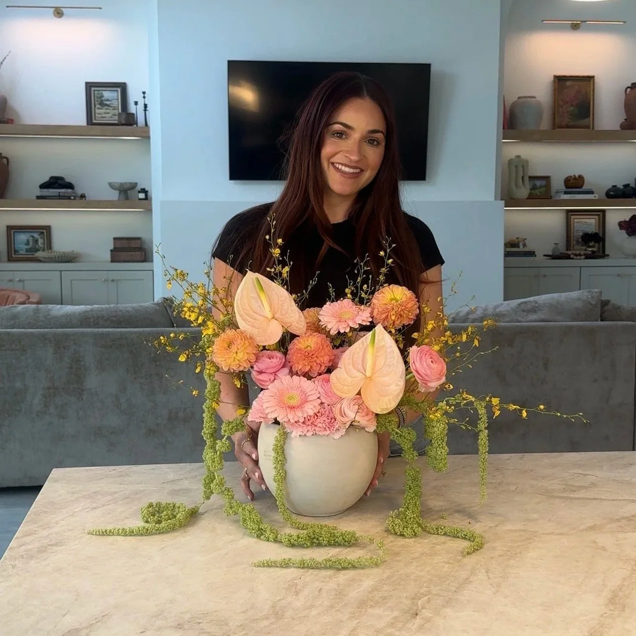 A woman with long dark hair smiling, sitting behind a large floral arrangement in a white vase on a beige table in a modern living room.