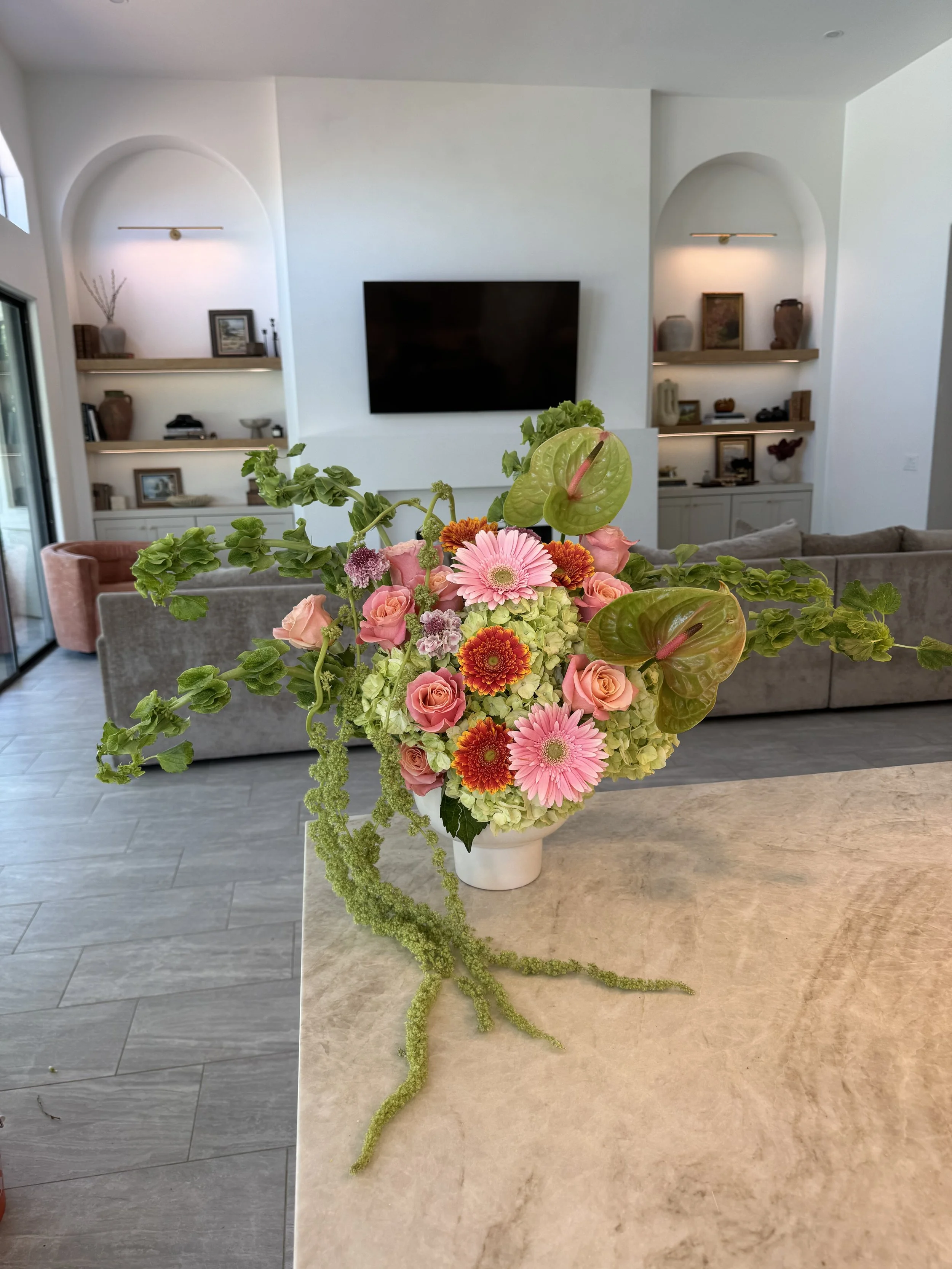 A colorful flower arrangement featuring pink roses, pink gerbera daisies, orange marigolds, green anthuriums, and lush green foliage in a white vase on a kitchen island.