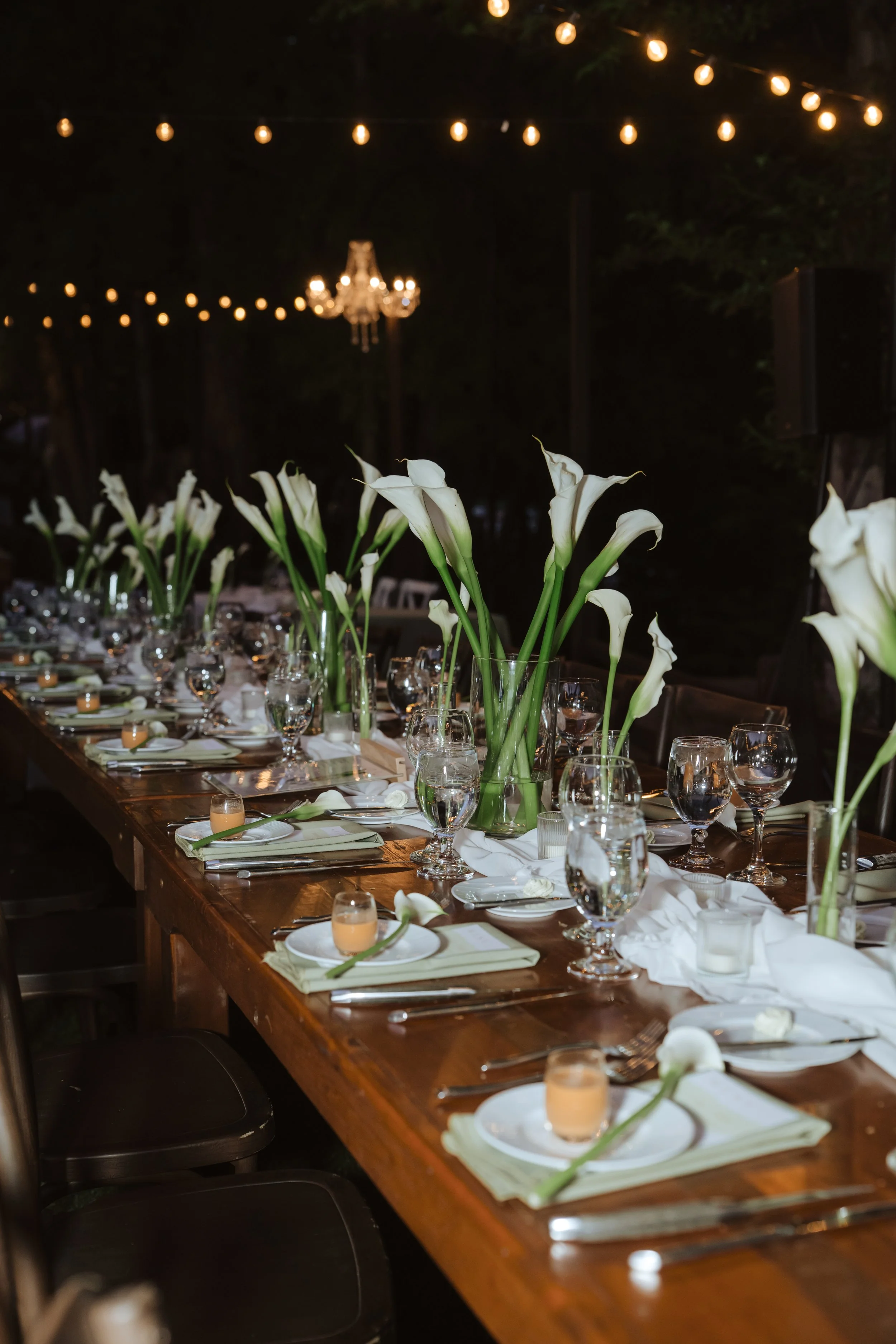 Nighttime outdoor dining setup with a long wooden table decorated with white calla lily flowers in glass vases, white napkins, and glassware, under hanging string lights and a chandelier in the background.