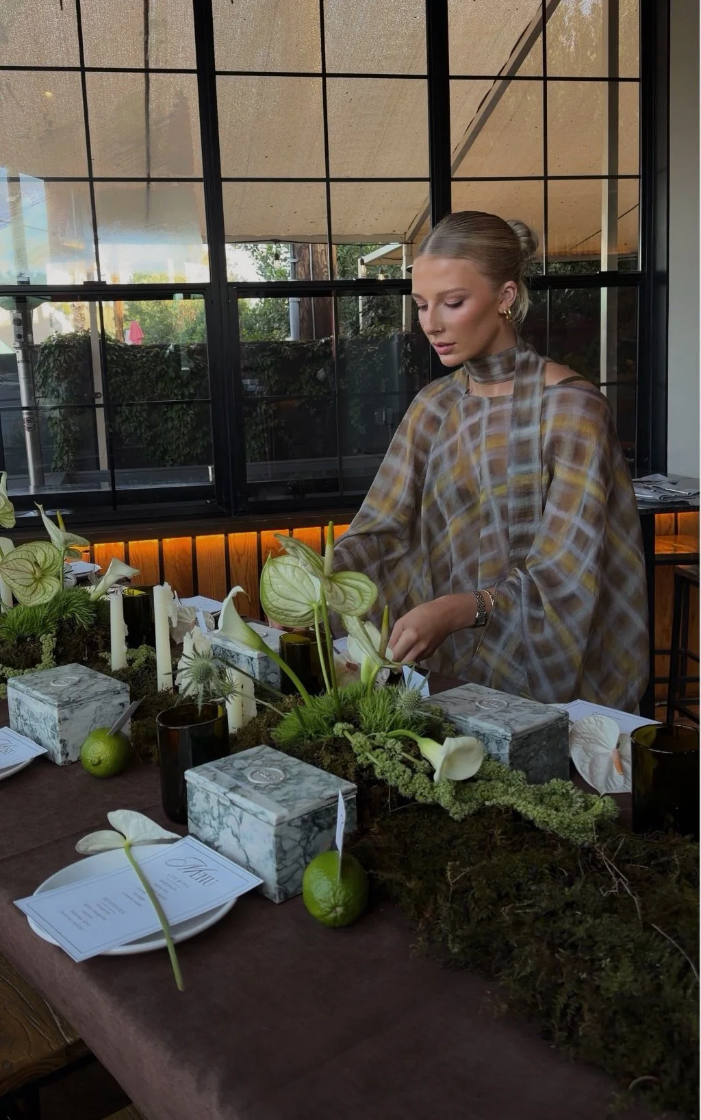 A woman with blonde hair tied in a bun, wearing a brown and beige plaid dress with a scarf around her neck, arranging flowers on a table decorated with greenery, white flowers, candles, marble boxes, and stationery, inside a room with large windows.