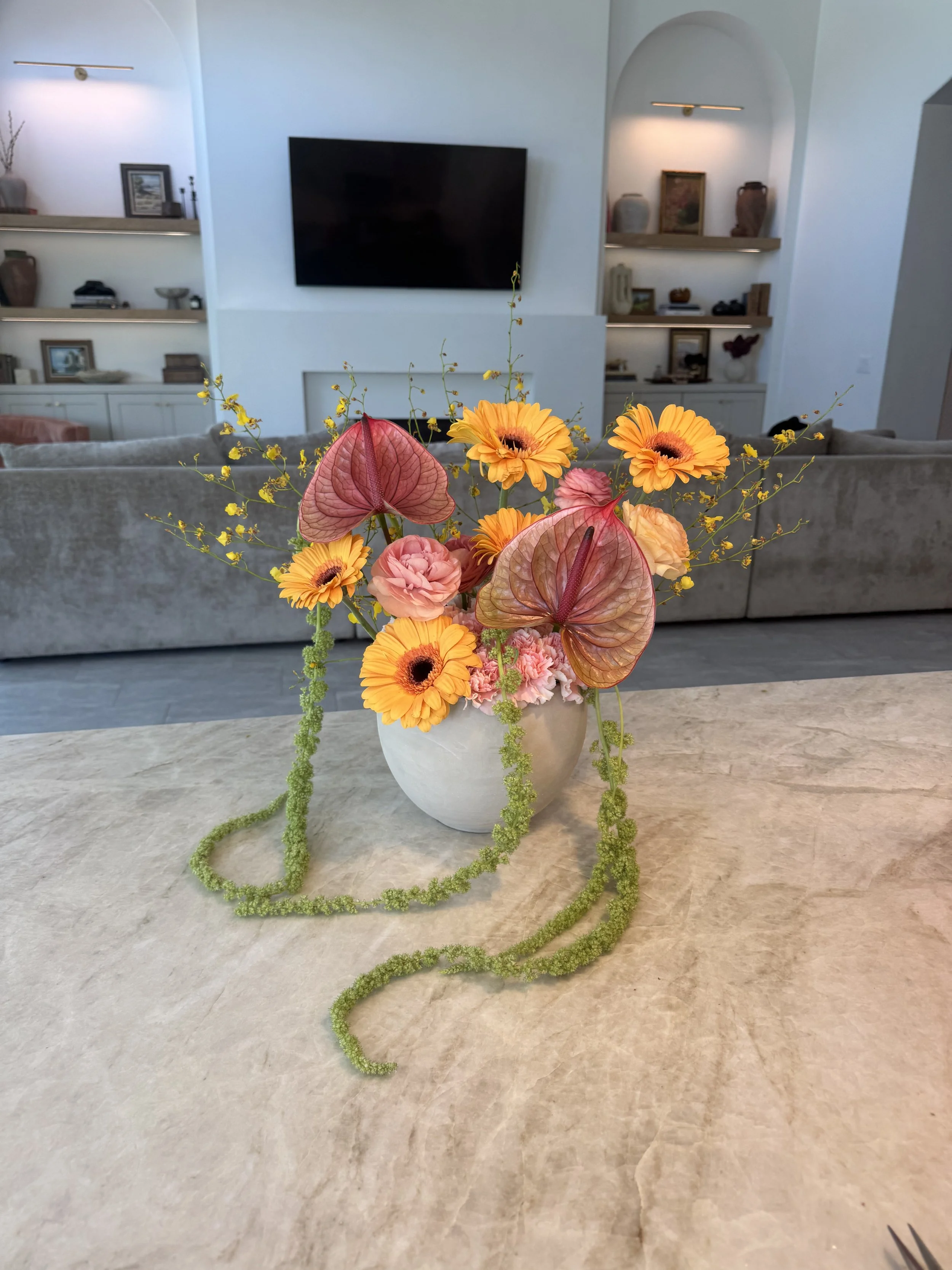 A vase with a colorful flower arrangement on a table in a modern living room, with a TV and built-in shelves in the background.