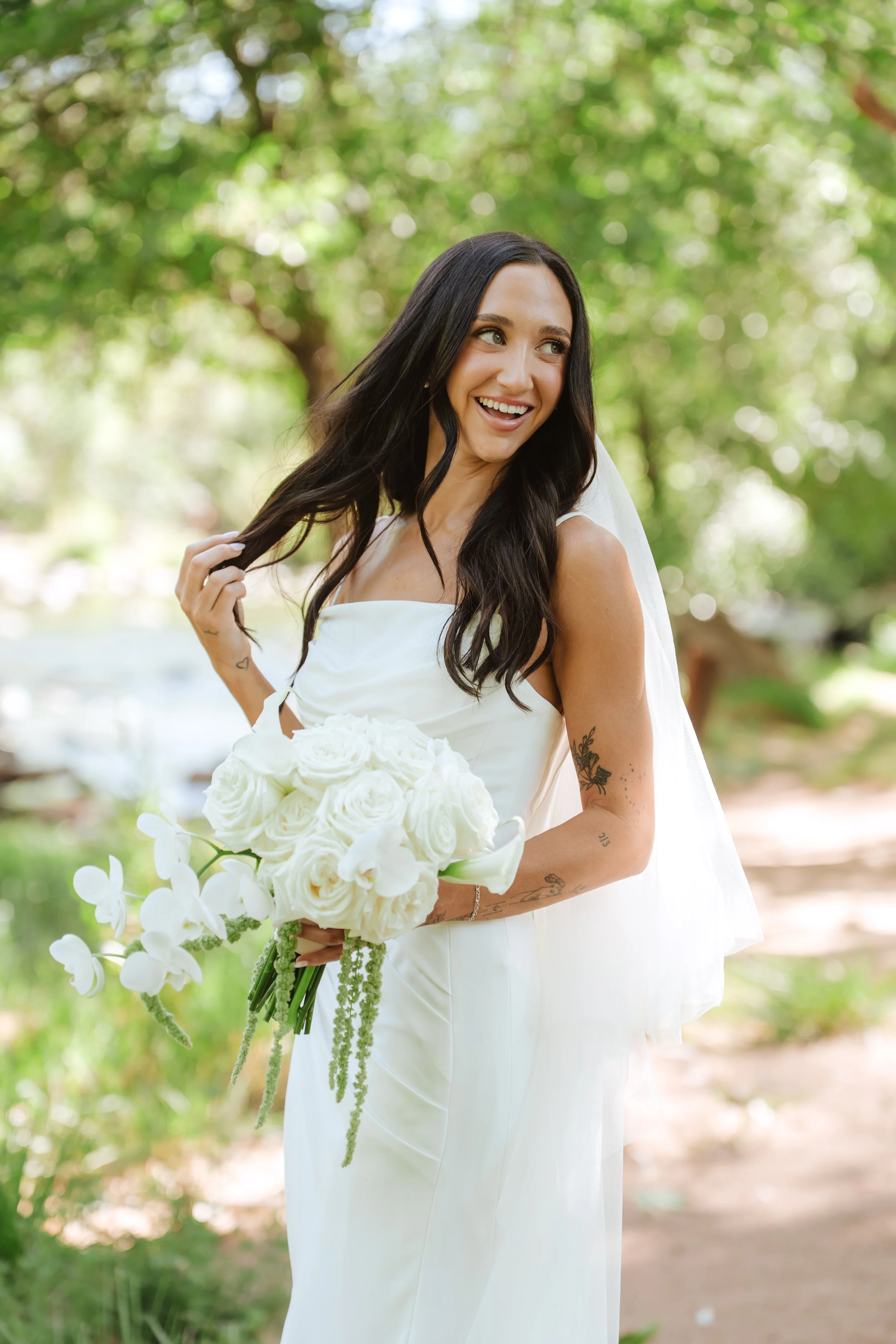 A woman in a white dress holding a bouquet of white flowers, smiling and looking to her right, outdoors with green trees in the background.