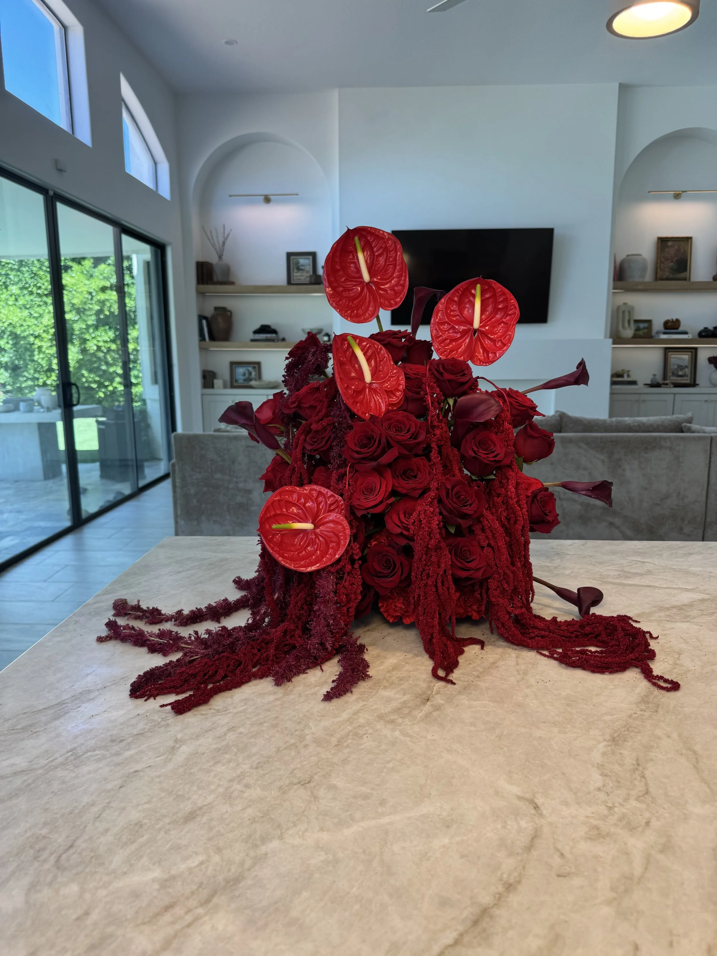 A floral arrangement with red roses, red anthuriums, and red amaranth on a marble table in a modern living room.