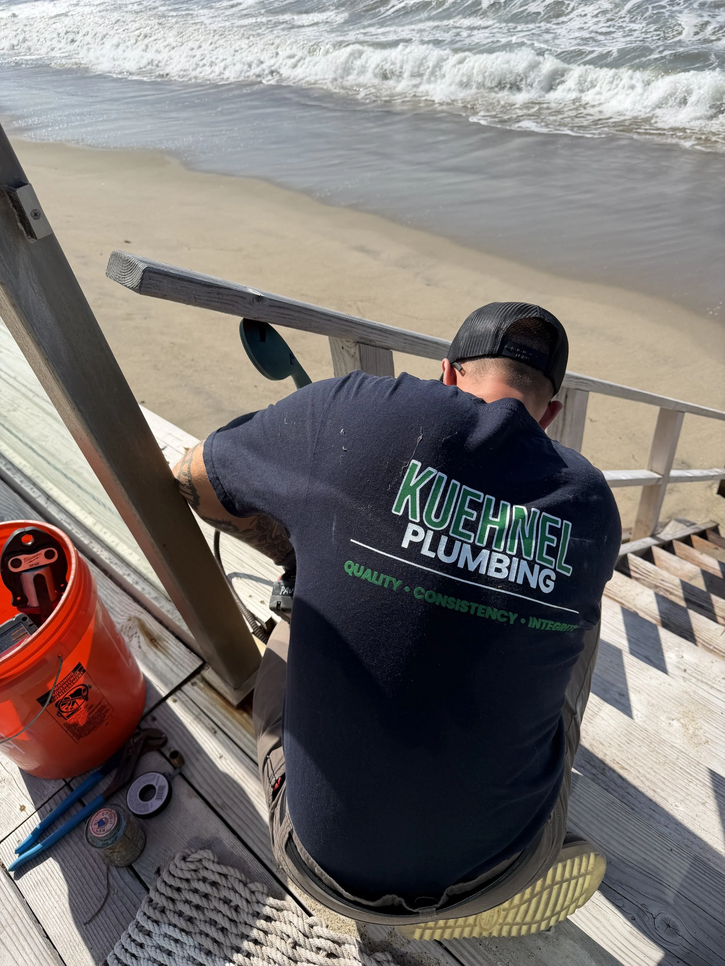 A man working on plumbing repairs on a wooden deck near the beach, with ocean waves in the background.