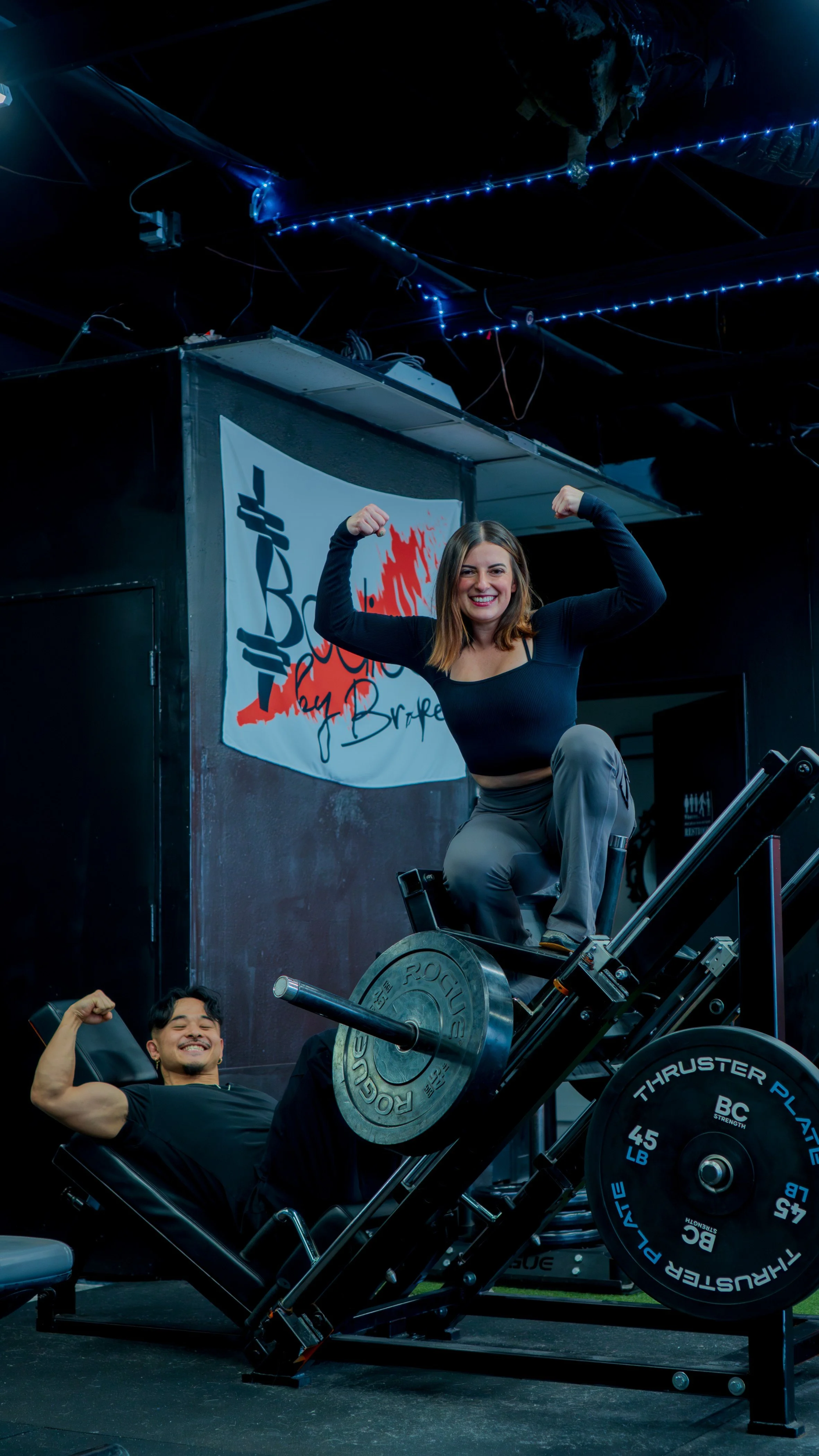 Two people at the gym, one woman on a leg press machine flexing her arms and smiling, a man lying on the machine smiling and flexing his arm, all in a dark gym with blue string lights and a banner with a medical symbol and red maple leaf in the backg