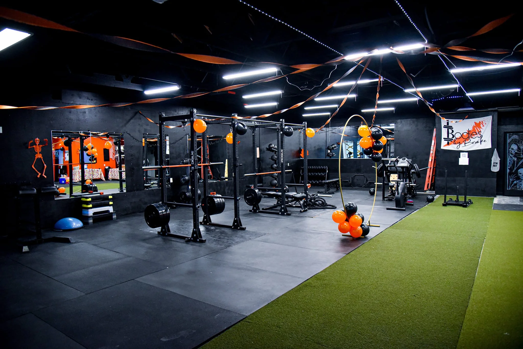 An indoor gym with black walls and ceiling, decorated with orange and black balloons and streamers. The gym has various workout equipment including weightlifting racks, barbells, a stationary bike, and balance balls. There is a green turf area on the right side and a wall mirror in the background.
