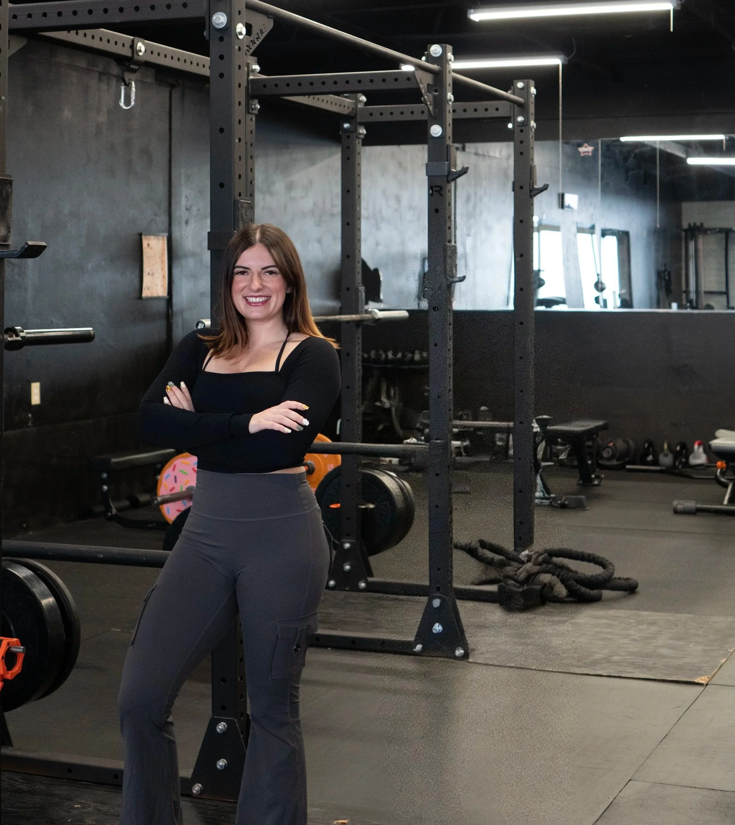 A woman standing in a gym with arms crossed, smiling, in front of weightlifting equipment and gym tools.