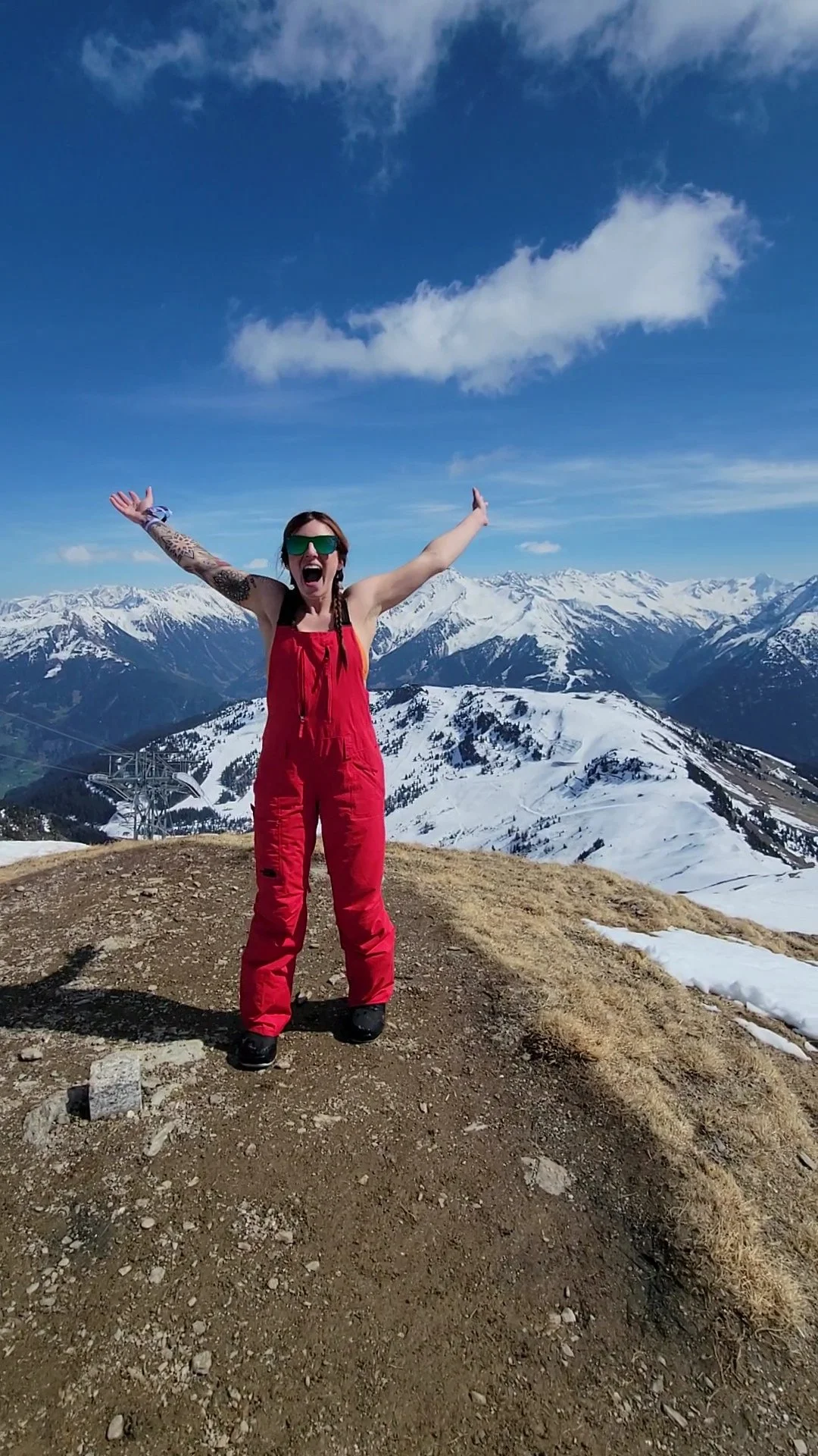 Woman in red ski suit celebrating on mountain top with snow-covered mountains and blue sky in the background.