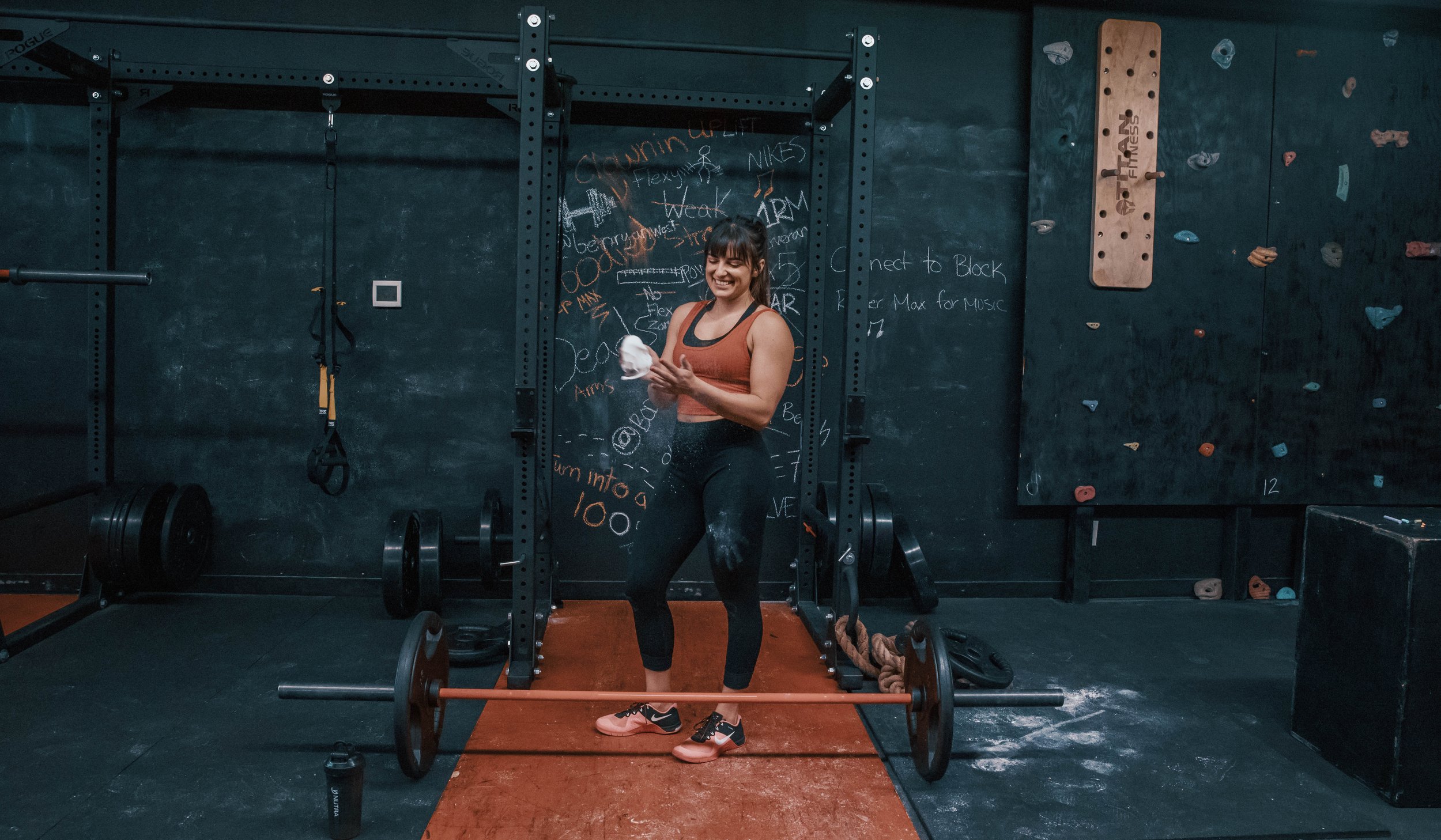 Woman in athletic wear smiling and wiping sweat in a gym, with weights, black chalkboard walls, and climbing holds in the background.