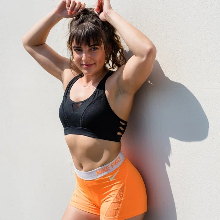 A young woman in workout attire standing against a plain white wall, smiling, with her hands in her hair.