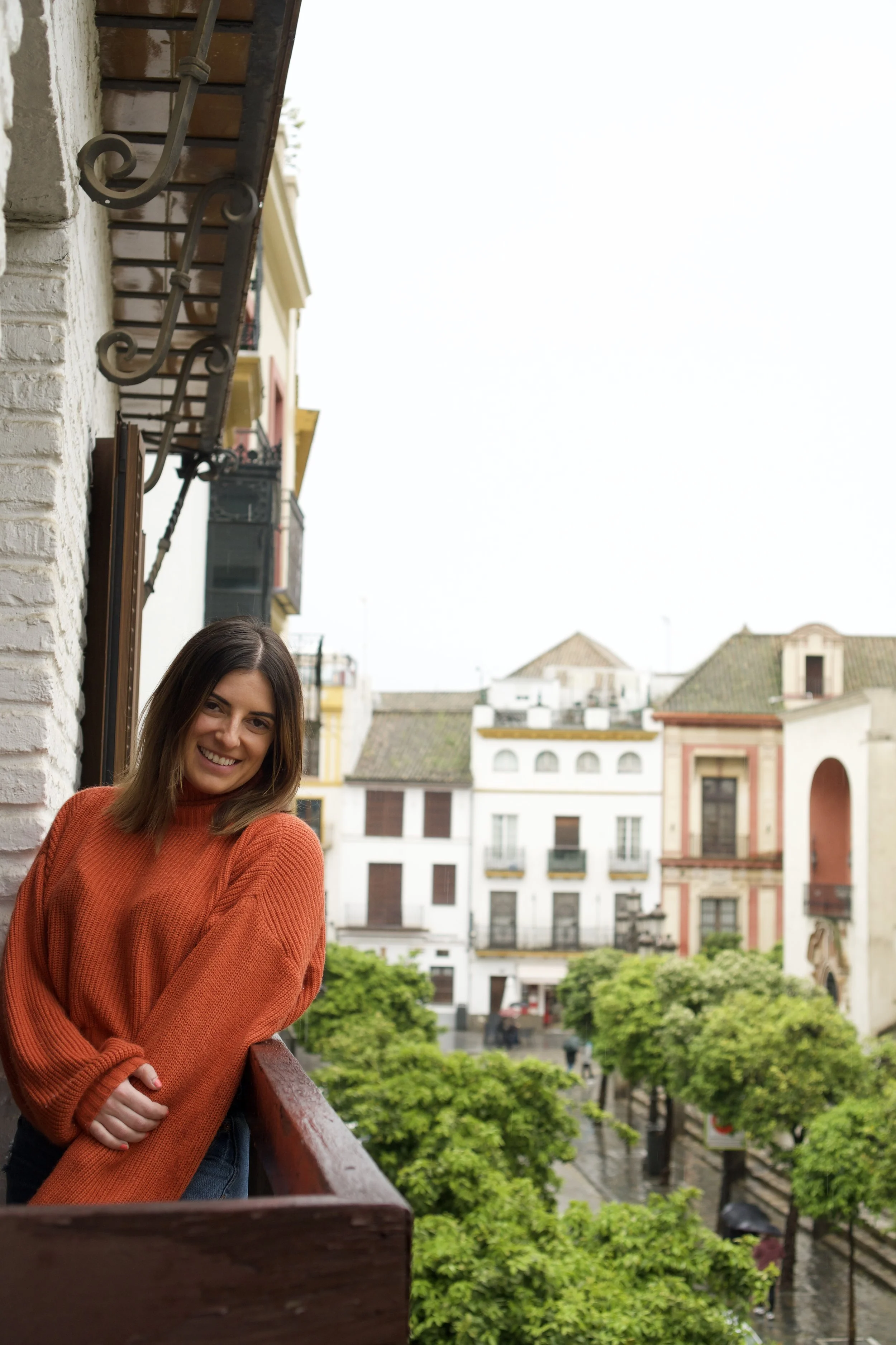 A young woman with shoulder-length brown hair smiles while leaning on a balcony railing in an urban area with trees and colonial-style buildings in the background.