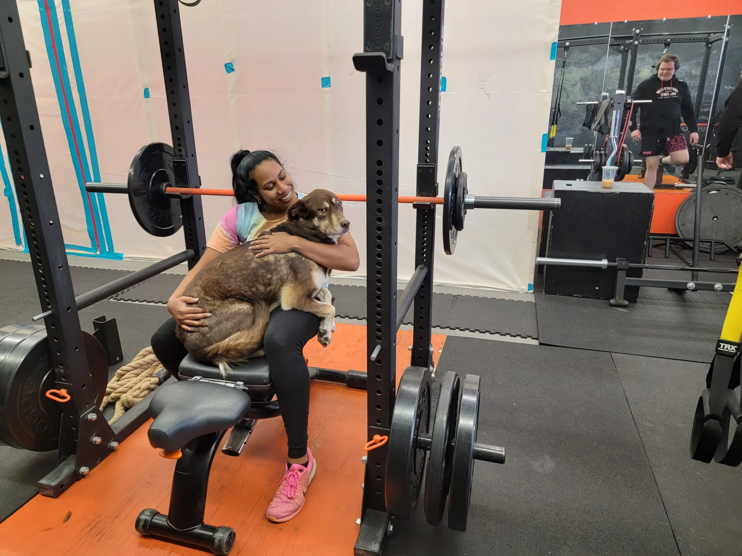 A woman is sitting on a bench in a gym, hugging a large, mixed-breed dog. The woman is smiling and wearing pink running shoes. The gym has black rubber flooring, weightlifting equipment, and a mirror in the background reflecting other gym members.