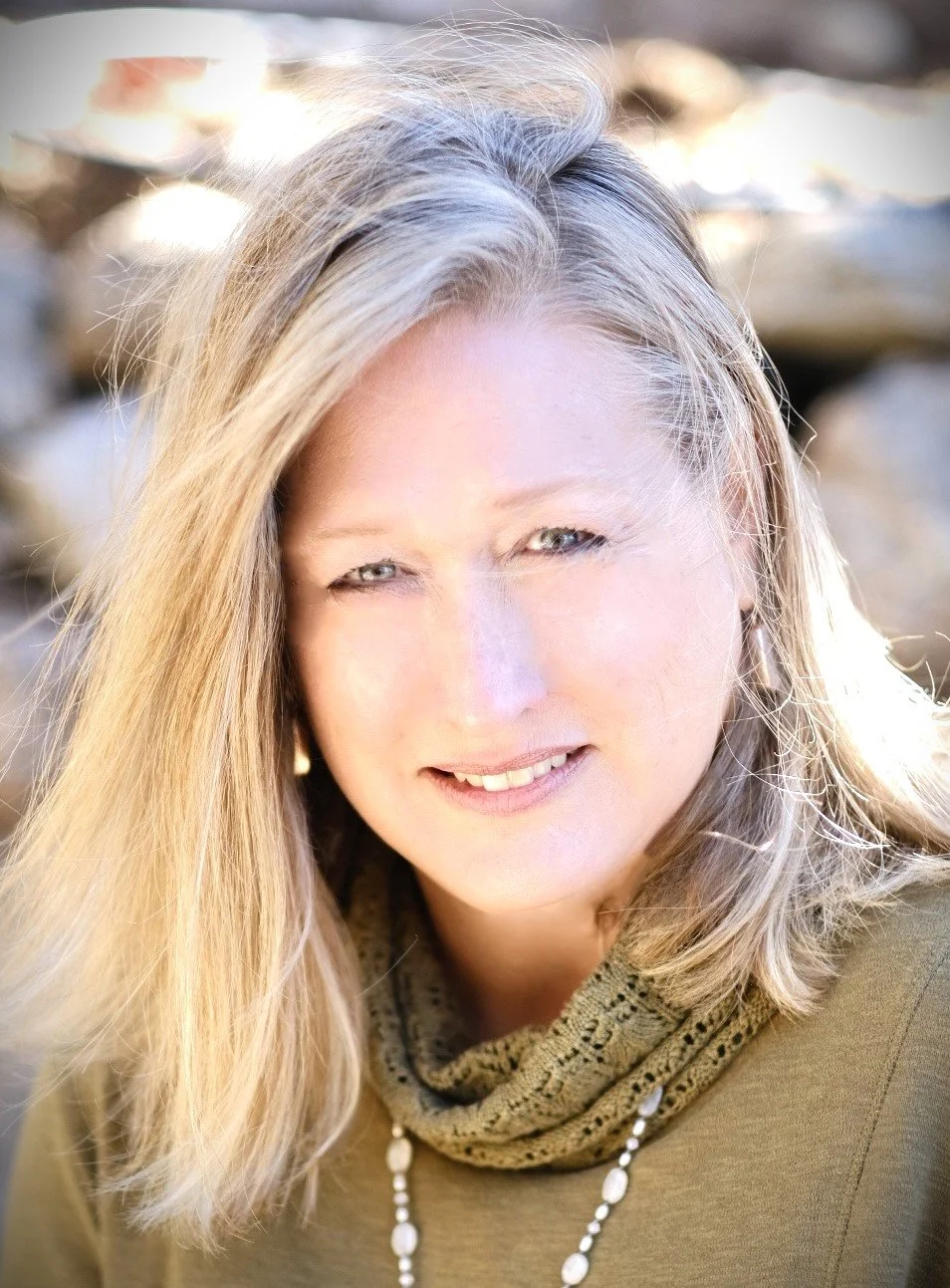 A woman with blonde hair smiling outdoors, wearing a scarf and necklace, with rocks in the background.