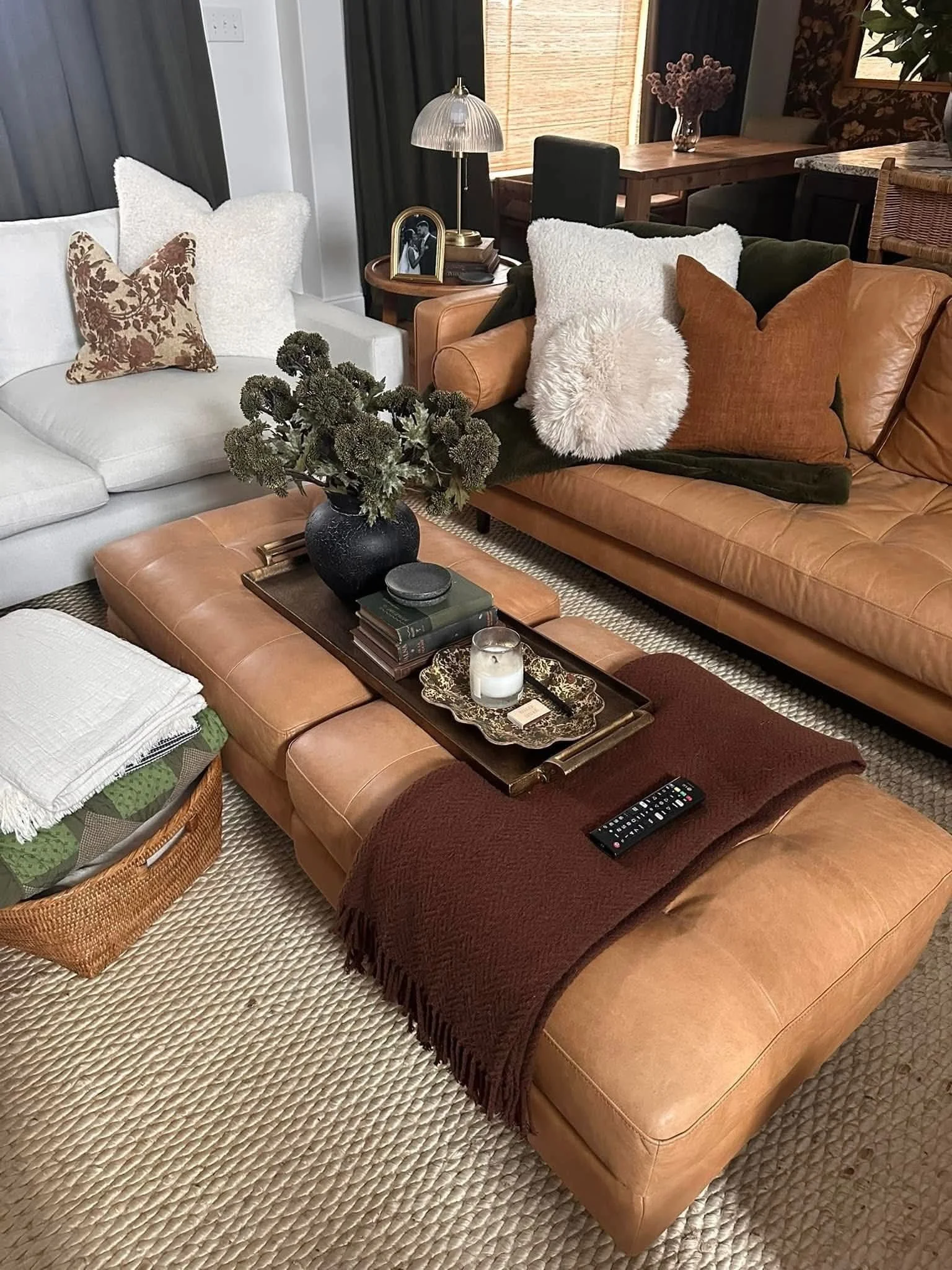 Living room with two sofas, a leather sofa with pillows, a white sofa with pillows, a wooden coffee table with a black vase of dried flowers, books, a candle, a remote, and a tray. There is a basket with blankets, a side table with a framed photo and a lamp, and a dining area in the background with a table, chairs, and flower decor.