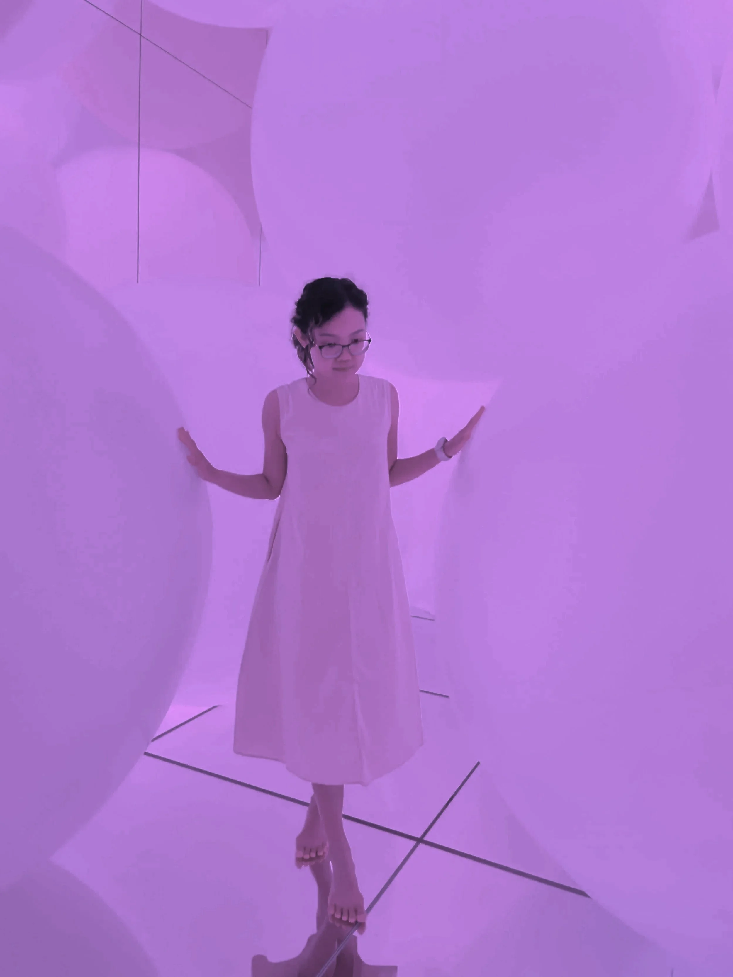 Person standing in an immersive artistic environment with purple lighting, large white spherical shapes, and curved reflective surfaces, wearing a sleeveless dress and glasses.