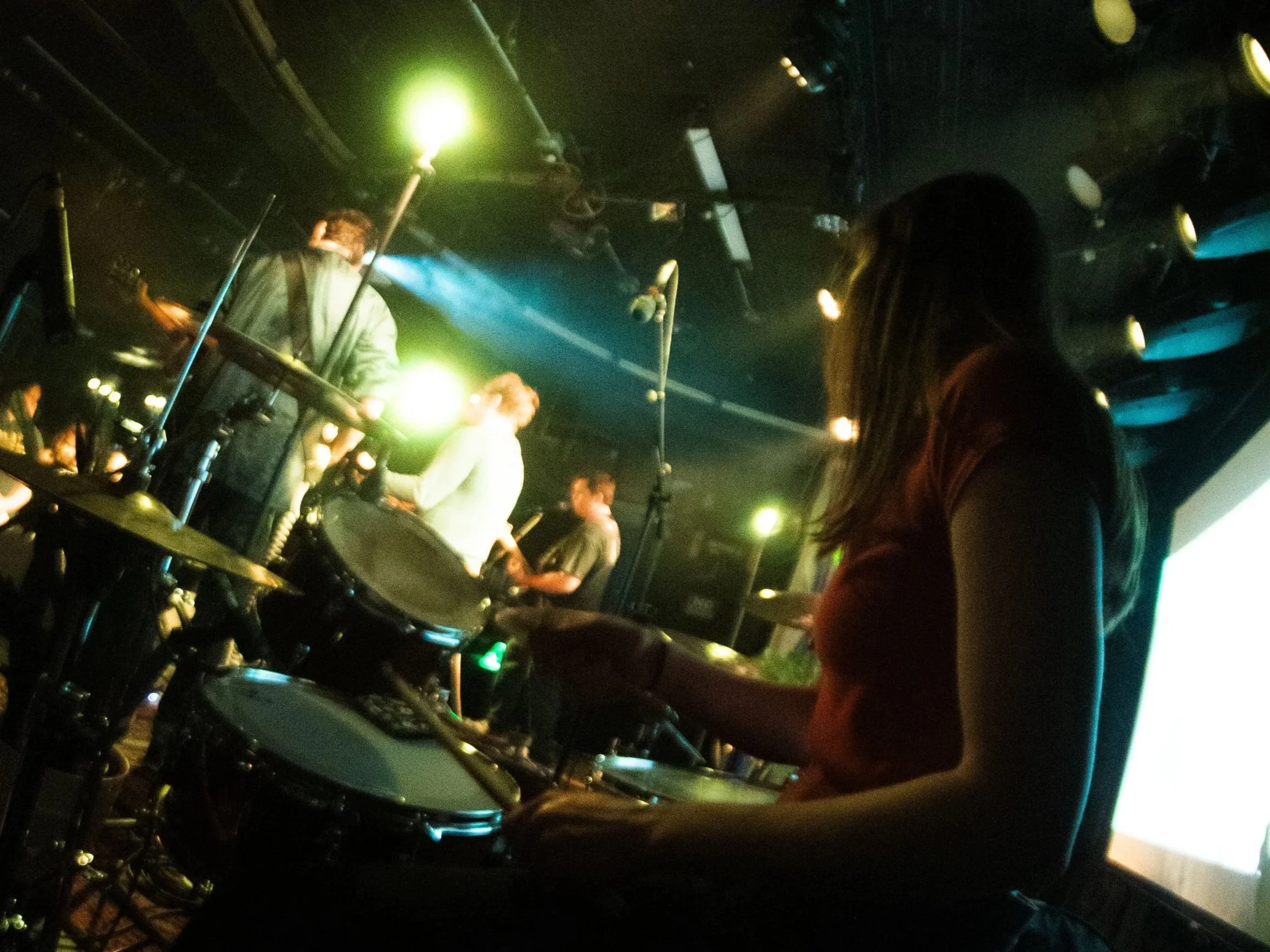 People performing music on stage in a dimly lit venue with green and blue stage lights.