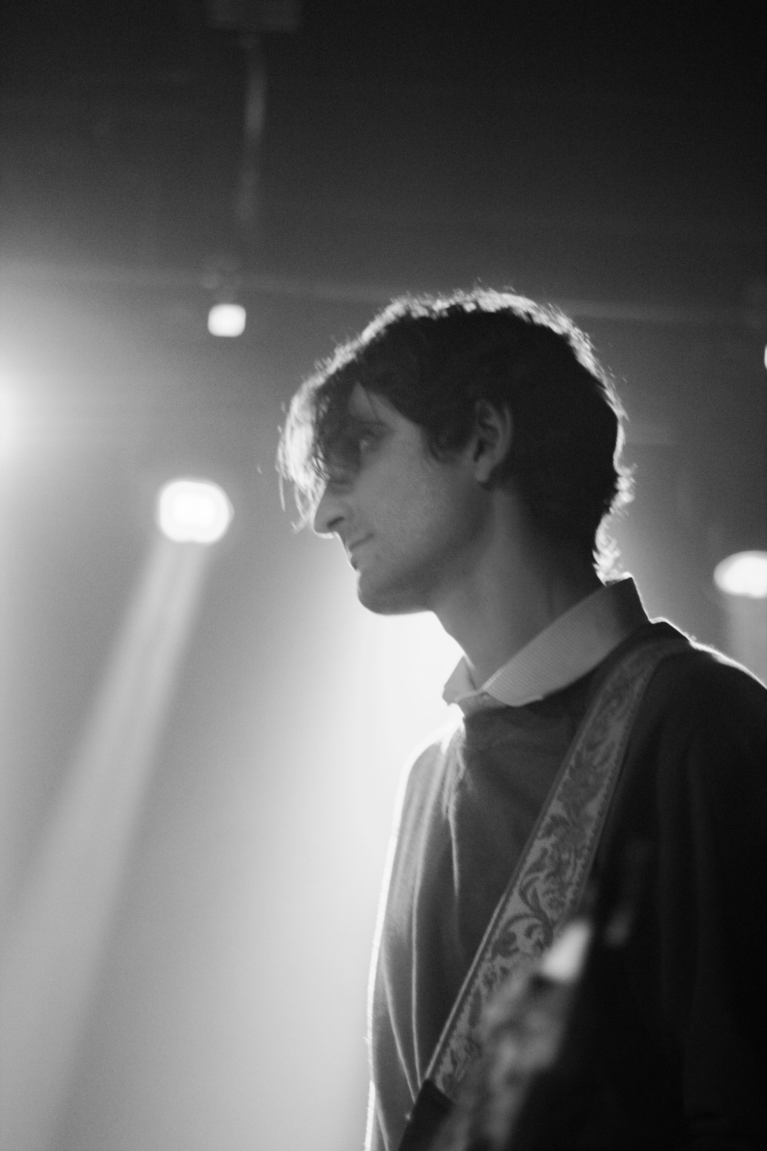 Side profile of a young man with dark hair, wearing a sweater and collared shirt, illuminated by stage lights in a concert setting.