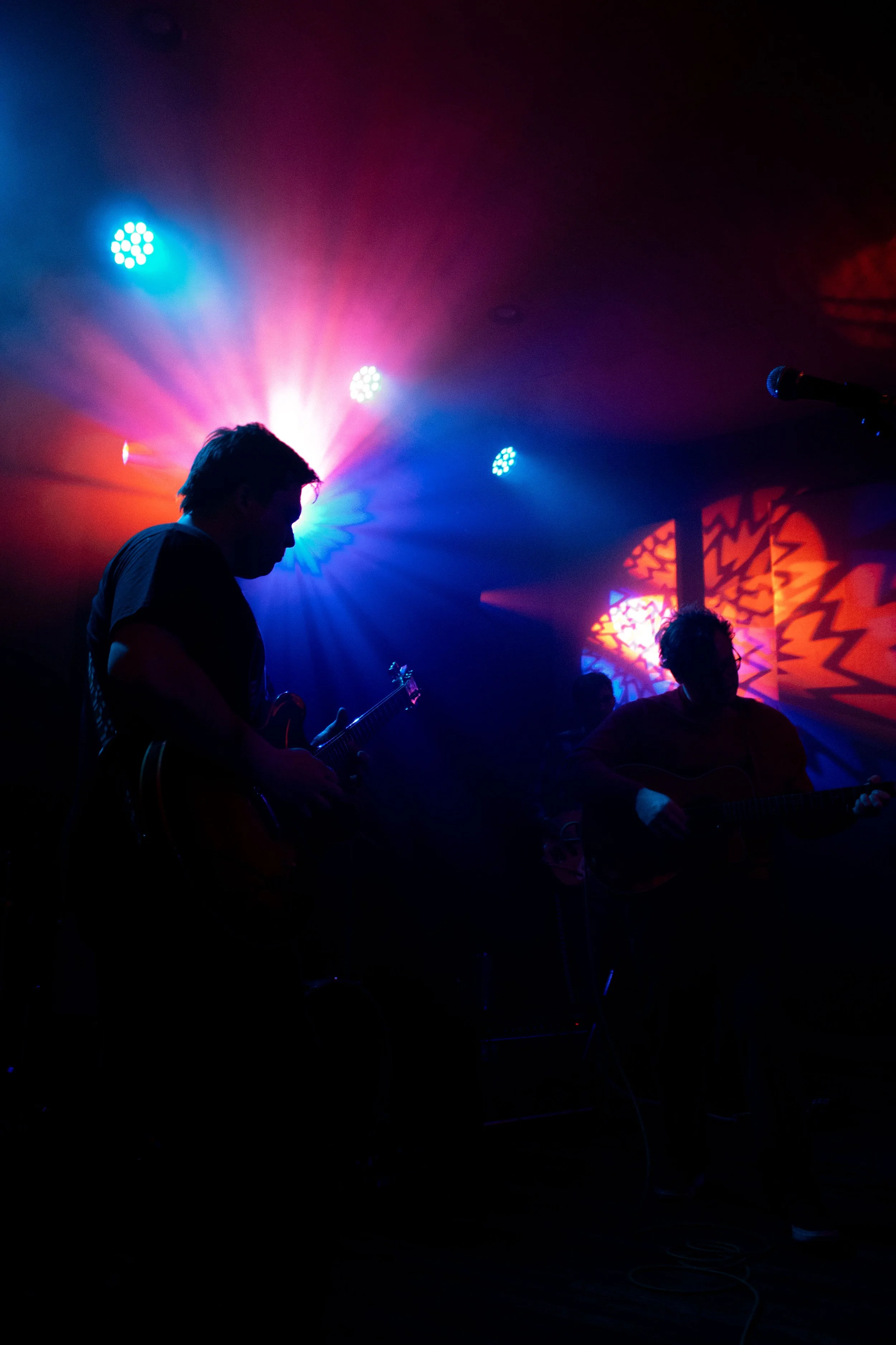 Silhouettes of musicians playing guitars on stage with vibrant colorful stage lights and patterns in the background.