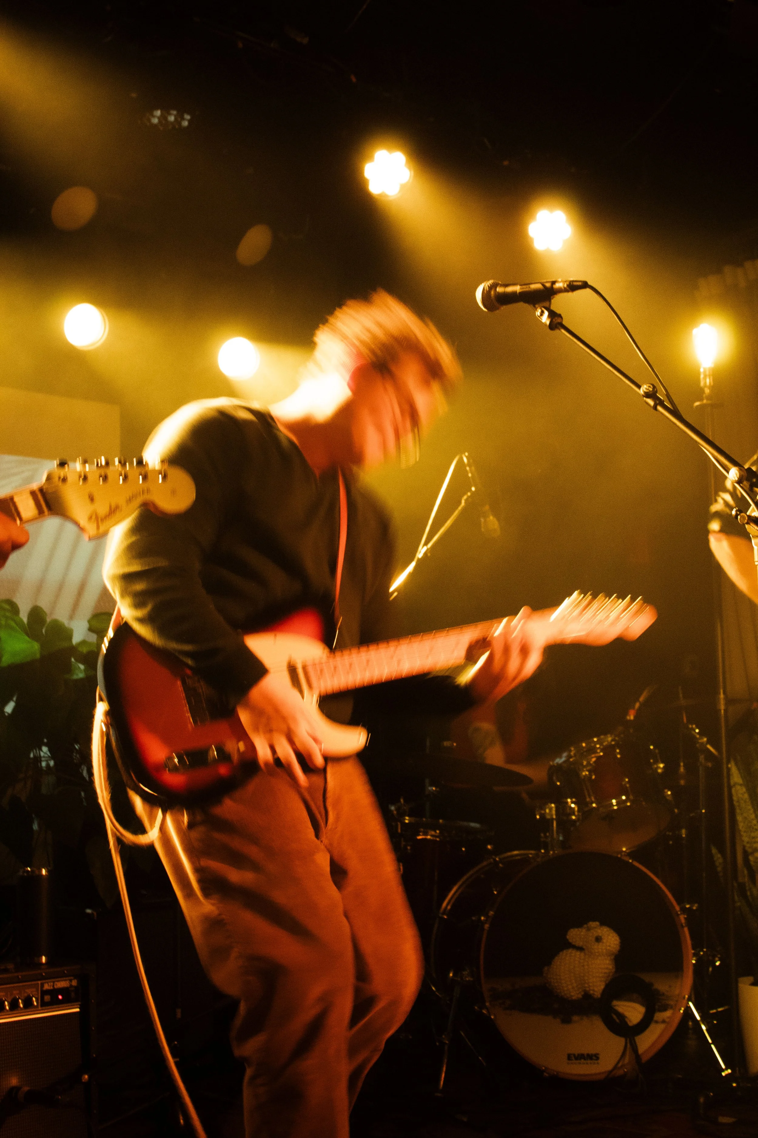 A musician playing an electric guitar on stage, with colorful stage lights and a drum set in the background.