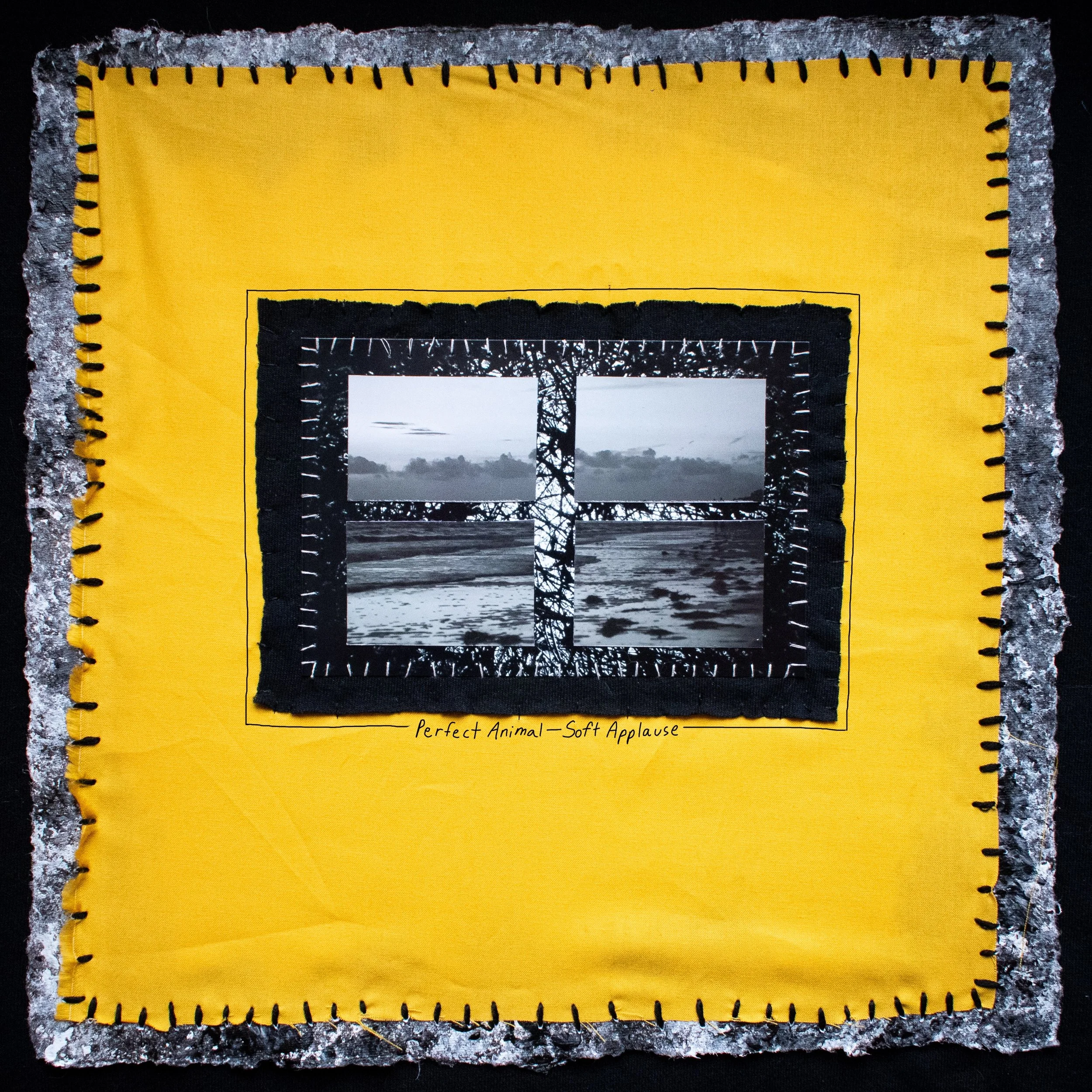 Mixed media artwork featuring a black and white photo collage of a beach scene with a tree silhouette, mounted on yellow fabric with decorative black stitching, and surrounded by textured gray material, with handwritten caption 'Perfect Animal - Soft Applause'.