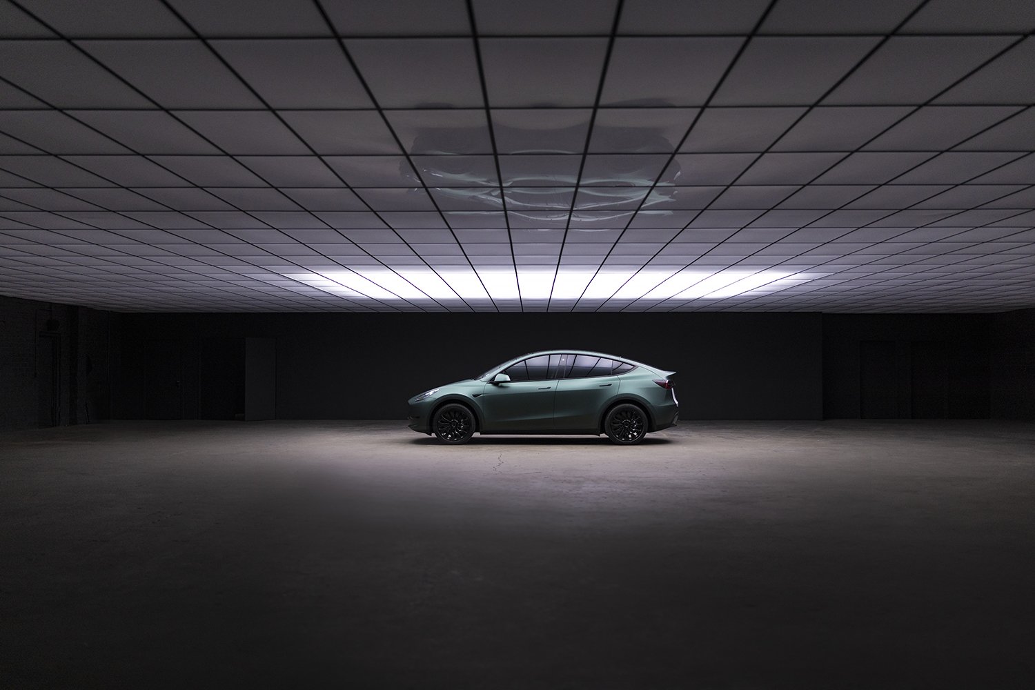 A dark indoor space with a single modern green SUV parked on a large concrete floor. The ceiling has a grid pattern with bright horizontal lighting and a reflective circular detail.