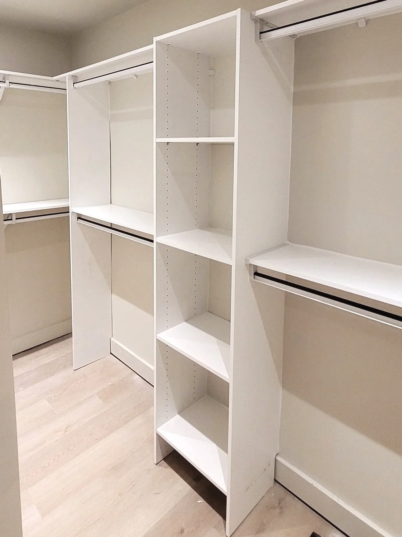 Empty walk-in closet with white shelving and hanging rods, hardwood floors, and beige walls.