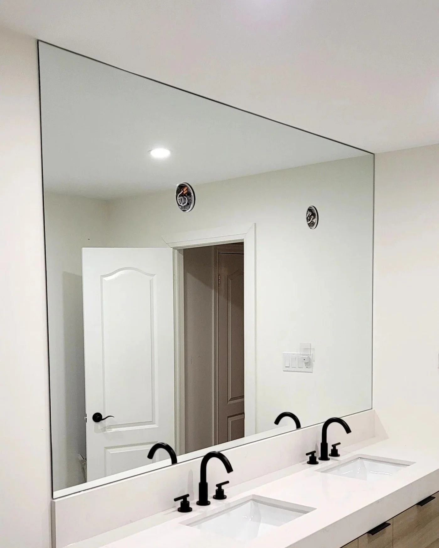 Bathroom with a large mirror above a double sink vanity, two black faucets, white countertop, white door, and two ceiling light fixtures. Some electrical outlets are visible.