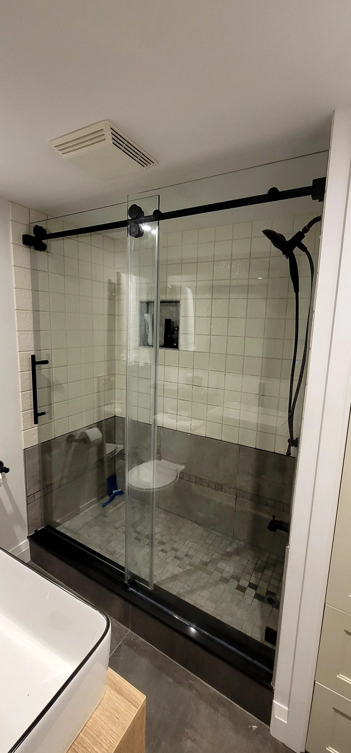A modern bathroom with a glass enclosed shower featuring black hardware, a mounted handheld shower head, white and gray tiles, a small recessed shelf, a white toilet, and a vent on the ceiling.