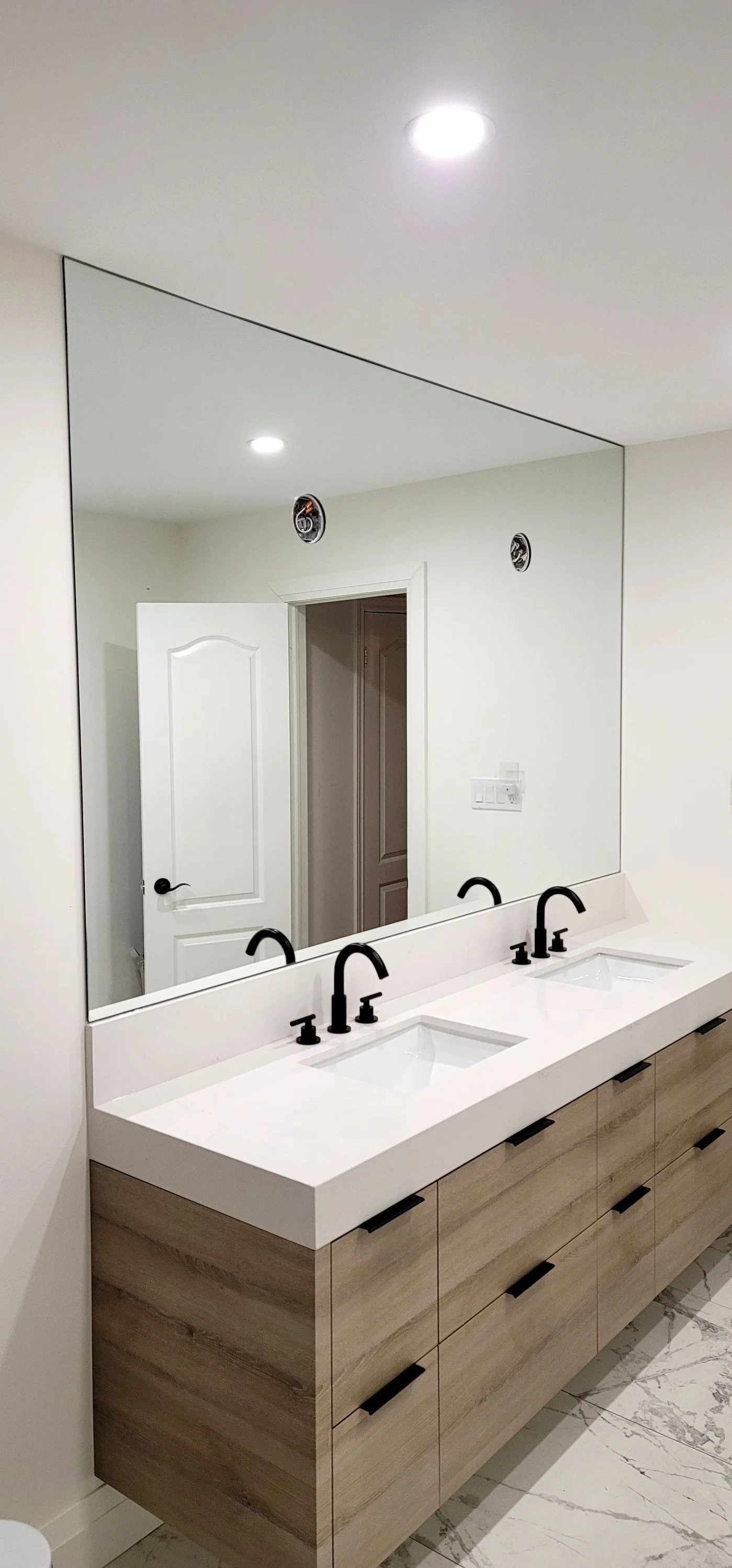 Modern bathroom vanity with a large mirror, three black faucets, and a wooden cabinet with black handles.