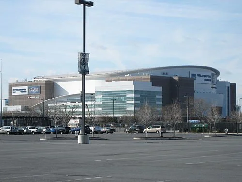 A large modern sports arena in an urban area with parking lot in front, daytime with clear sky.