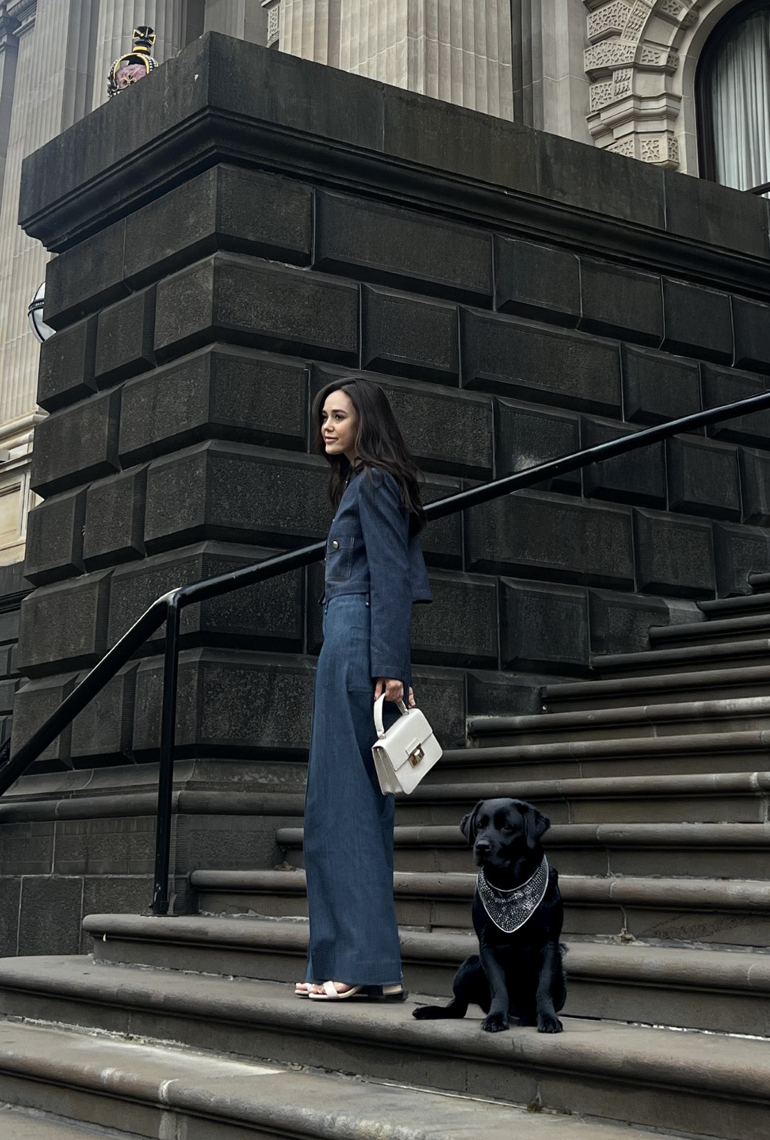 A woman in a blue denim pantsuit holding a white handbag stands on steps outside a historic building, with a black dog sitting beside her wearing a bandana.