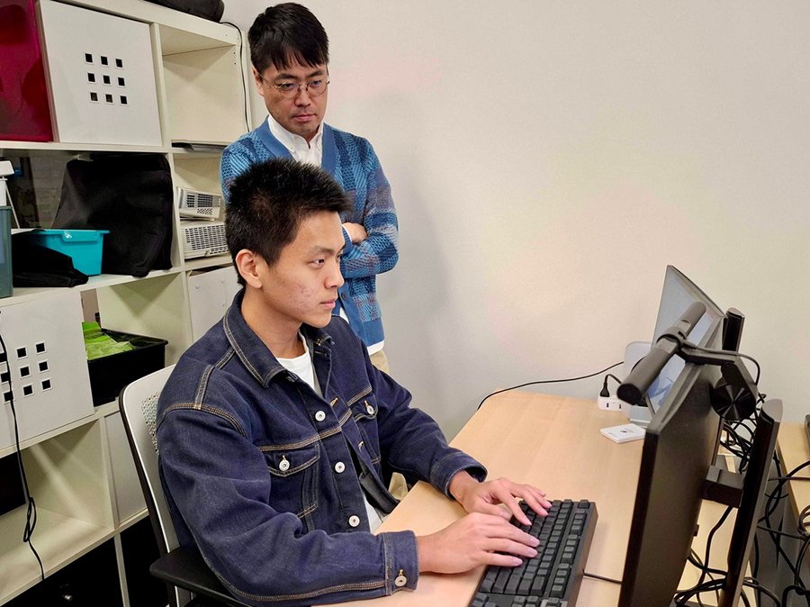 A student typing on a computer while a teacher looks over his shoulder