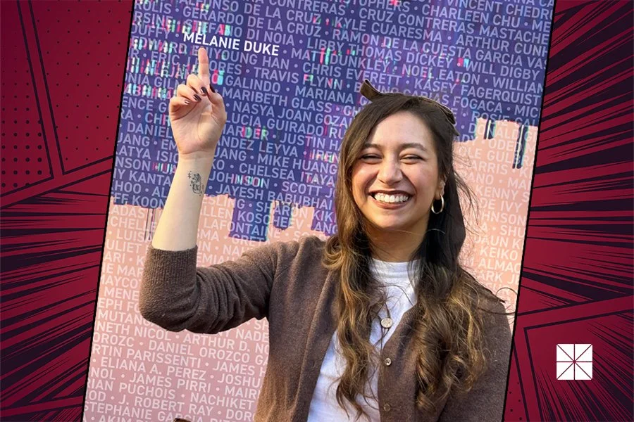 A young woman with long brown hair, wearing a brown cardigan and hoop earrings, is smiling and raising her right arm with her index finger pointing up. She is standing in front of a digital-looking backdrop filled with names and a colorful design.