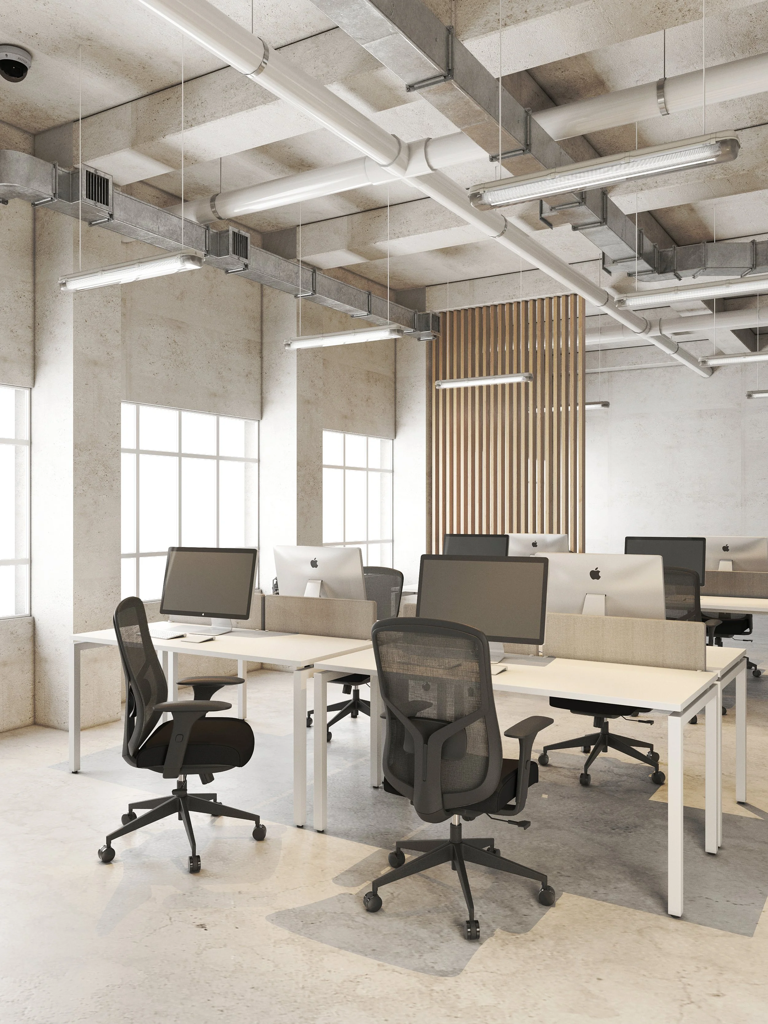 Oficina moderna con escritorios blancos, sillas ergonómicas negras, computadoras iMac en un espacio con ventanas grandes, paredes de concreto y techo industrial con tuberías y luces largas.