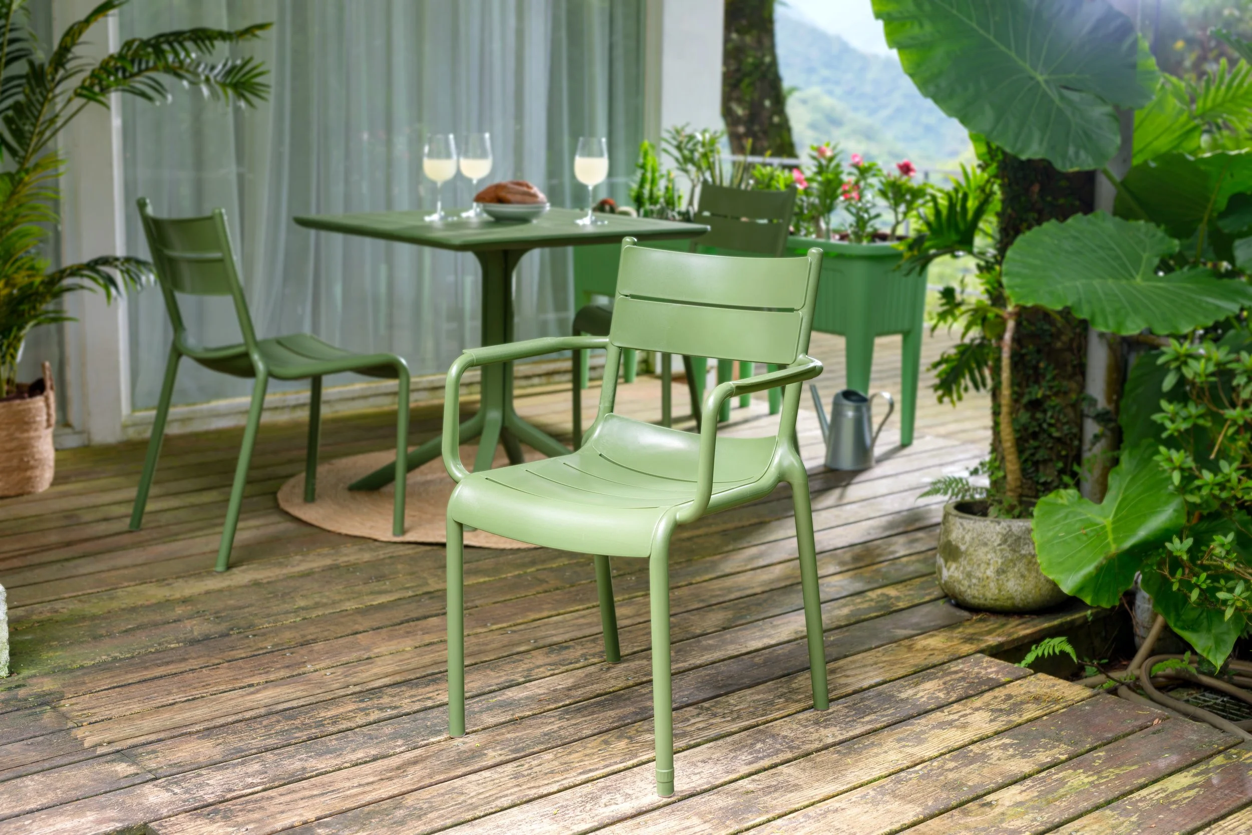 Silla de jardín de color verde en una terraza de madera, con plantas grandes y plantas con flores en macetas verdes, vistas a un paisaje natural con árboles y montañas.