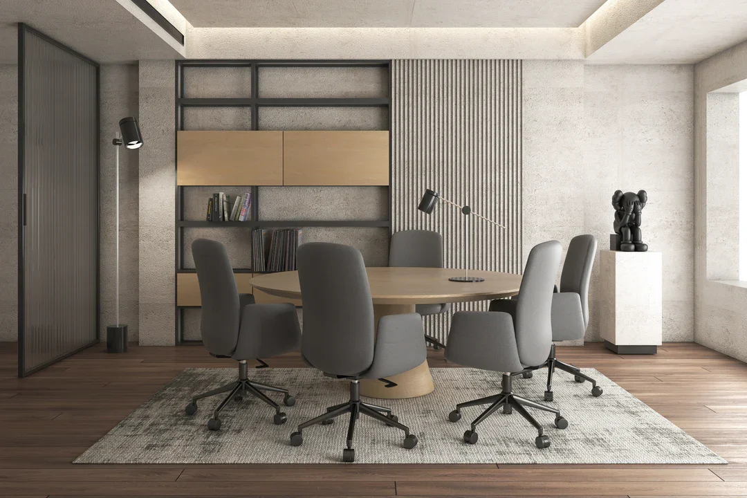 Sala de reuniones moderna con mesa ovalada de madera, ocho sillas con ruedas en gris, lámpara de escritorio negra, estatua decorativa en pedestal y estanterías con libros en fondo de pared de concreto.