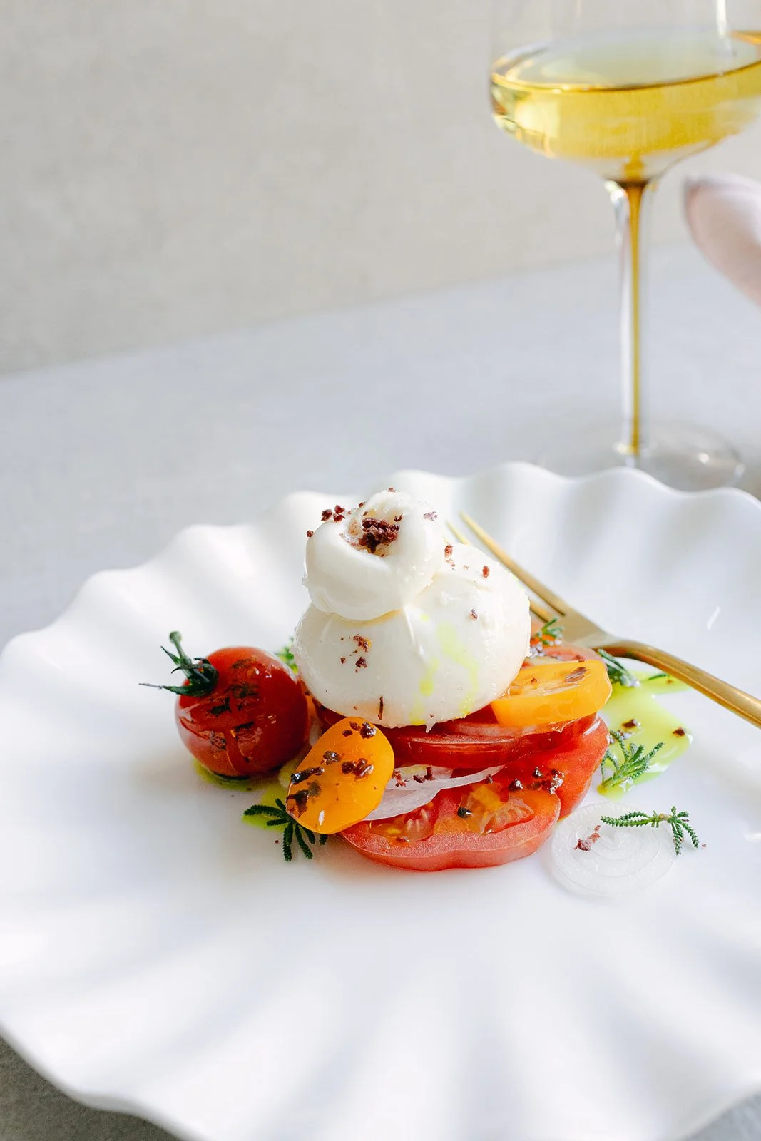 Caprese salad with heirloom tomatoes, mozzarella, cherry tomatoes, garnished with herbs, drizzled with olive oil, and accompanied by a glass of white wine.