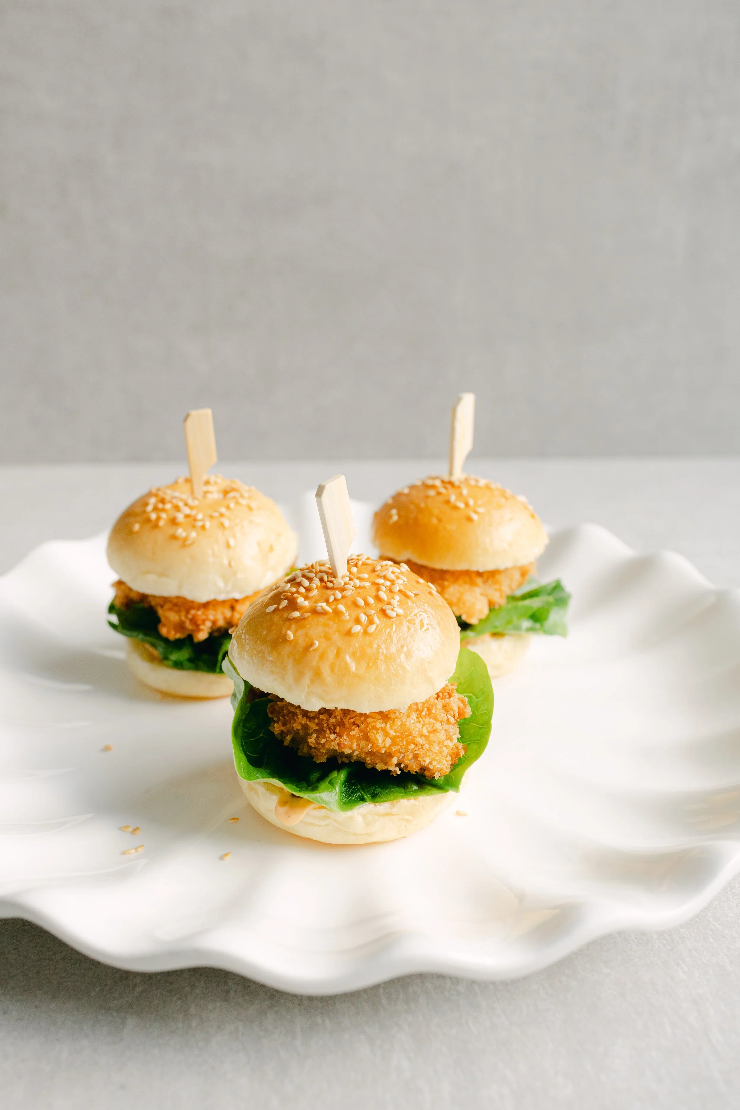 Mini chicken sandwiches with lettuce and sesame seed buns on a white decorative plate.