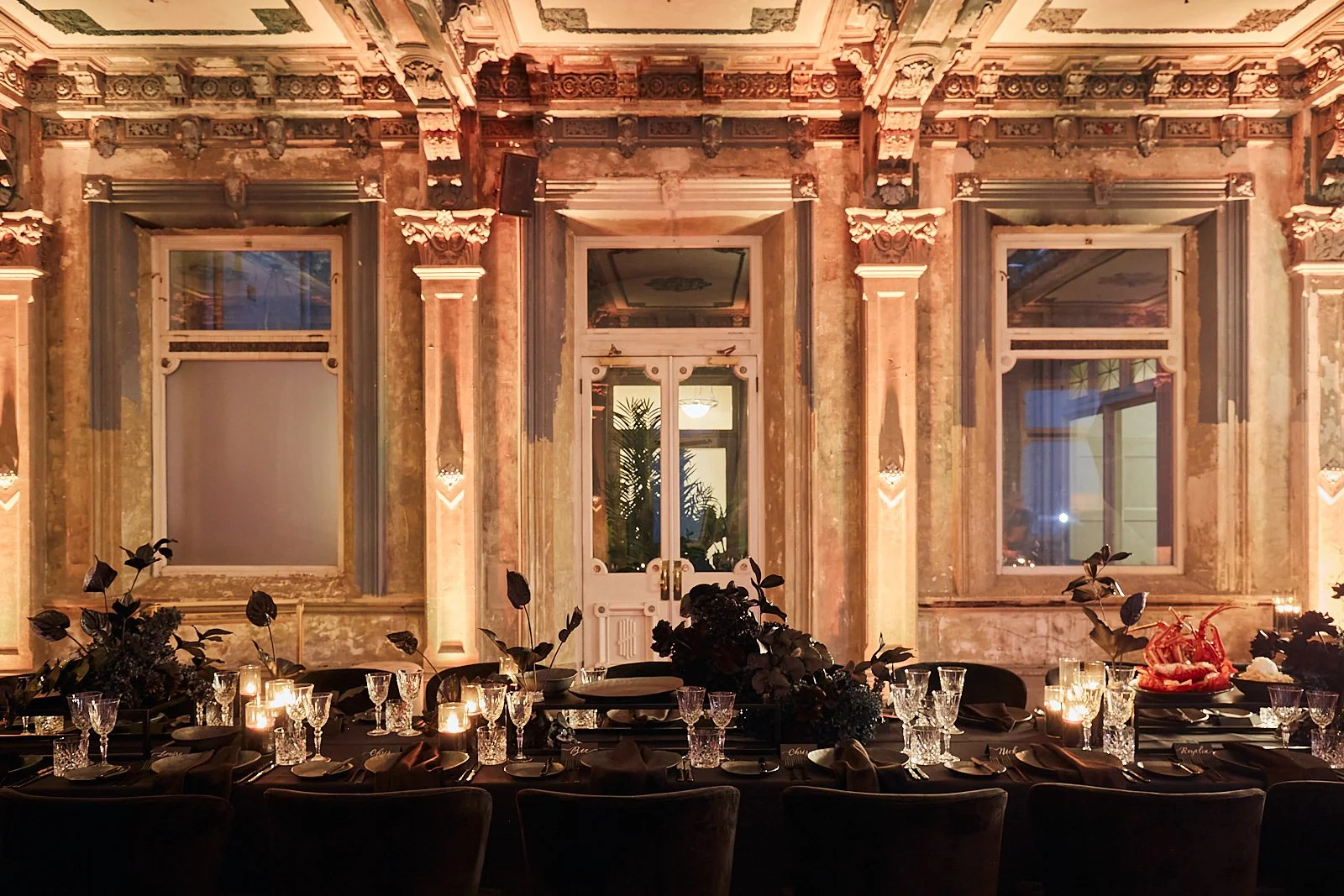 Elegant dining table set for a formal event in a spacious room with vintage decor, tall windows, and ornate molding, illuminated by candlelight.