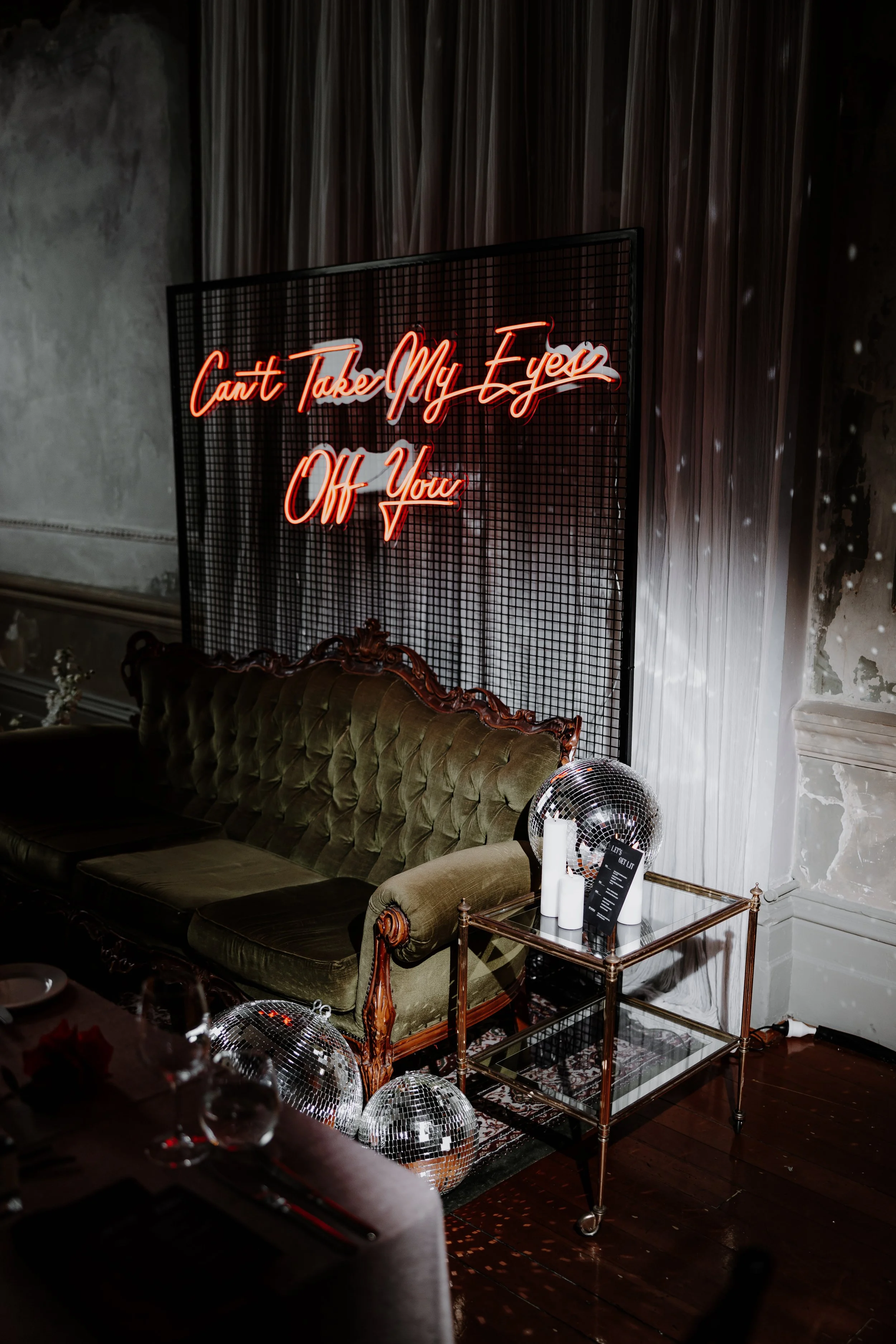 A neon sign reading 'Can't Take My Eyes Off You' behind a vintage green velvet sofa, with disco balls, candles, and a small glass table nearby in a dimly lit room with textured walls and curtains.