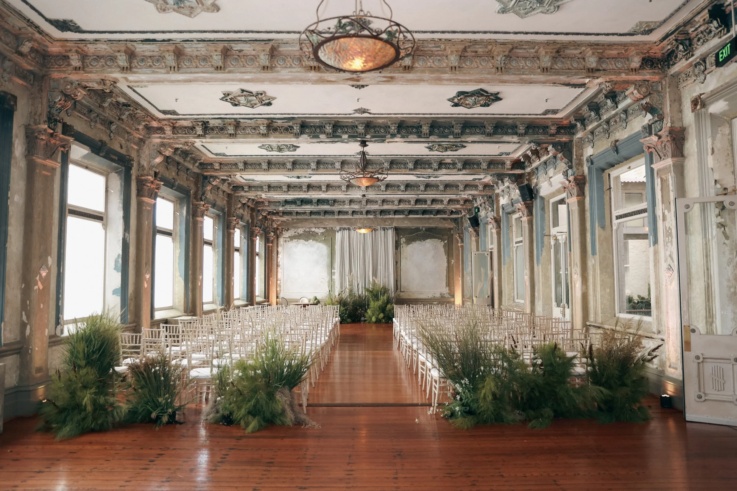 An ornate, vintage-style banquet hall with peeling paint and decorative moldings, set up for a wedding with white chairs arranged in rows along a wooden aisle, greenery and plants at the front, large windows on both sides, and a white curtain backdro