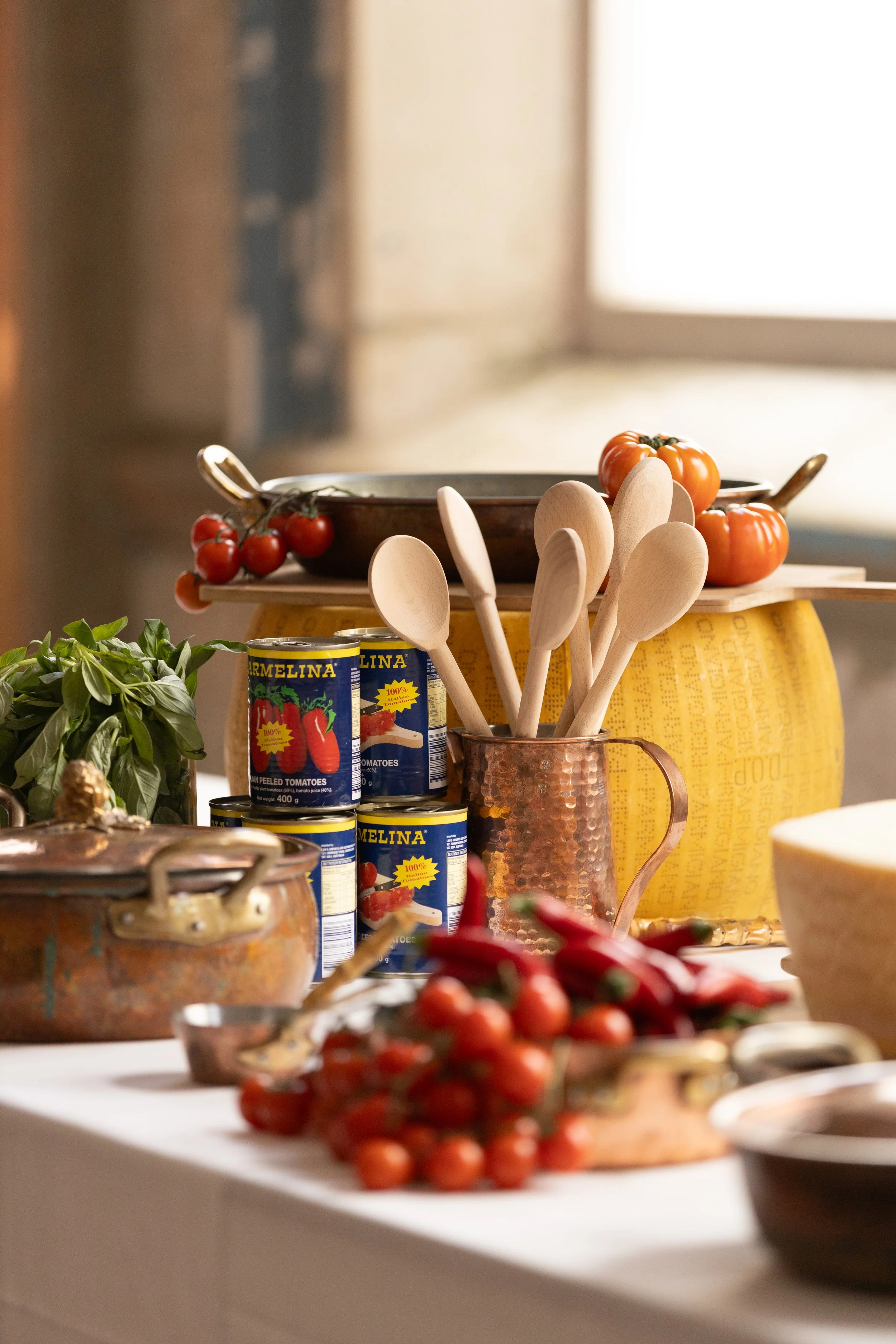 A kitchen counter with canned tomatoes, fresh cherry tomatoes, chili peppers, a copper container holding wooden spoons, a potted plant, and a yellow storage container.