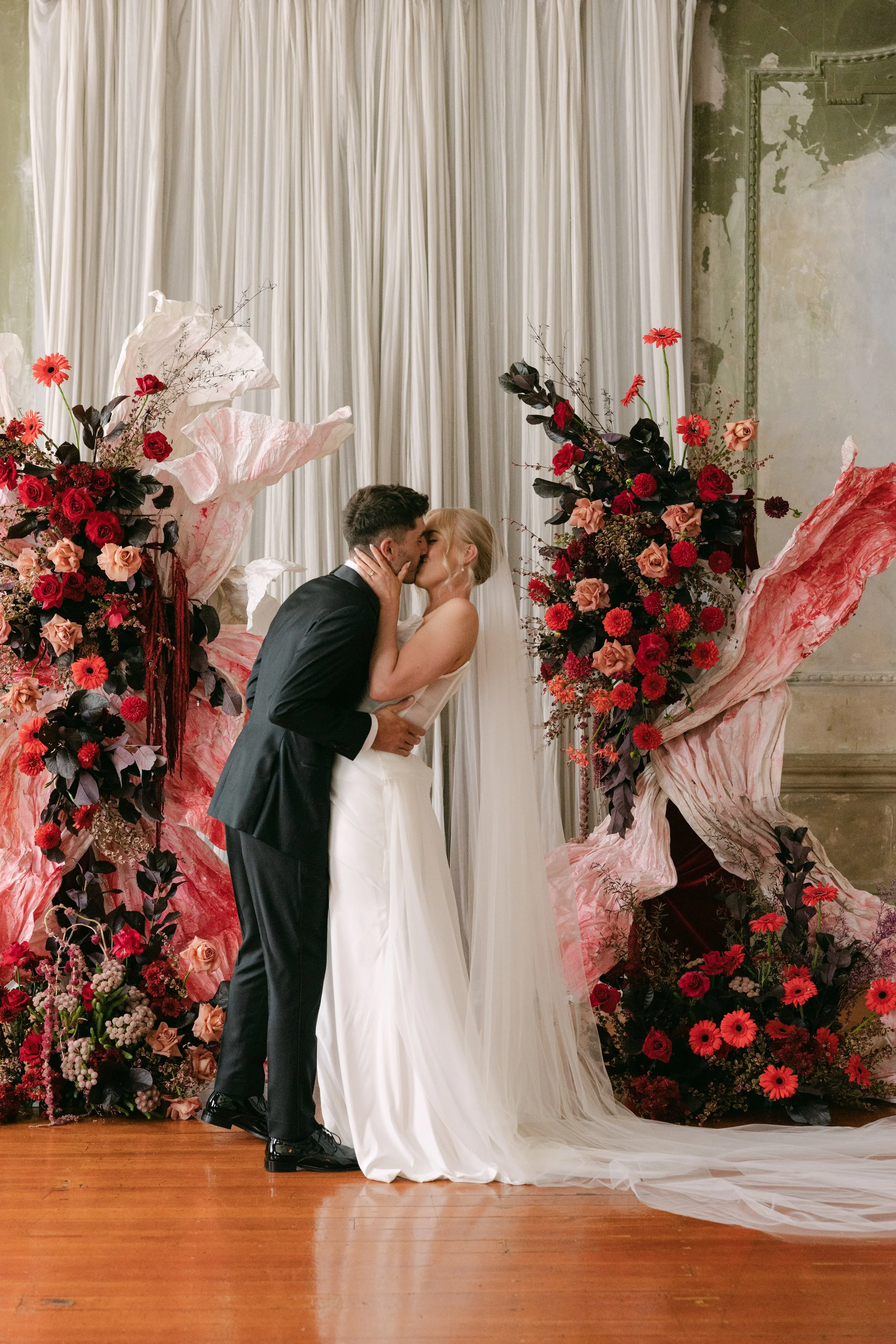A bride and groom sharing a kiss during their wedding, standing in front of floral arrangements with pink and red flowers and white curtains.